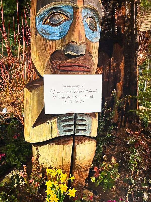 A wooden totem stands in a garden, commemorating Lieutenant Fred Obakow with a sign.