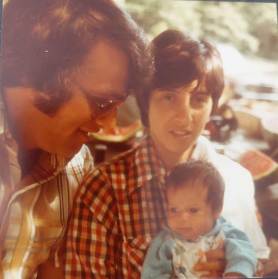 A joyful couple holds their baby, smiling warmly at each other in a sunny park.