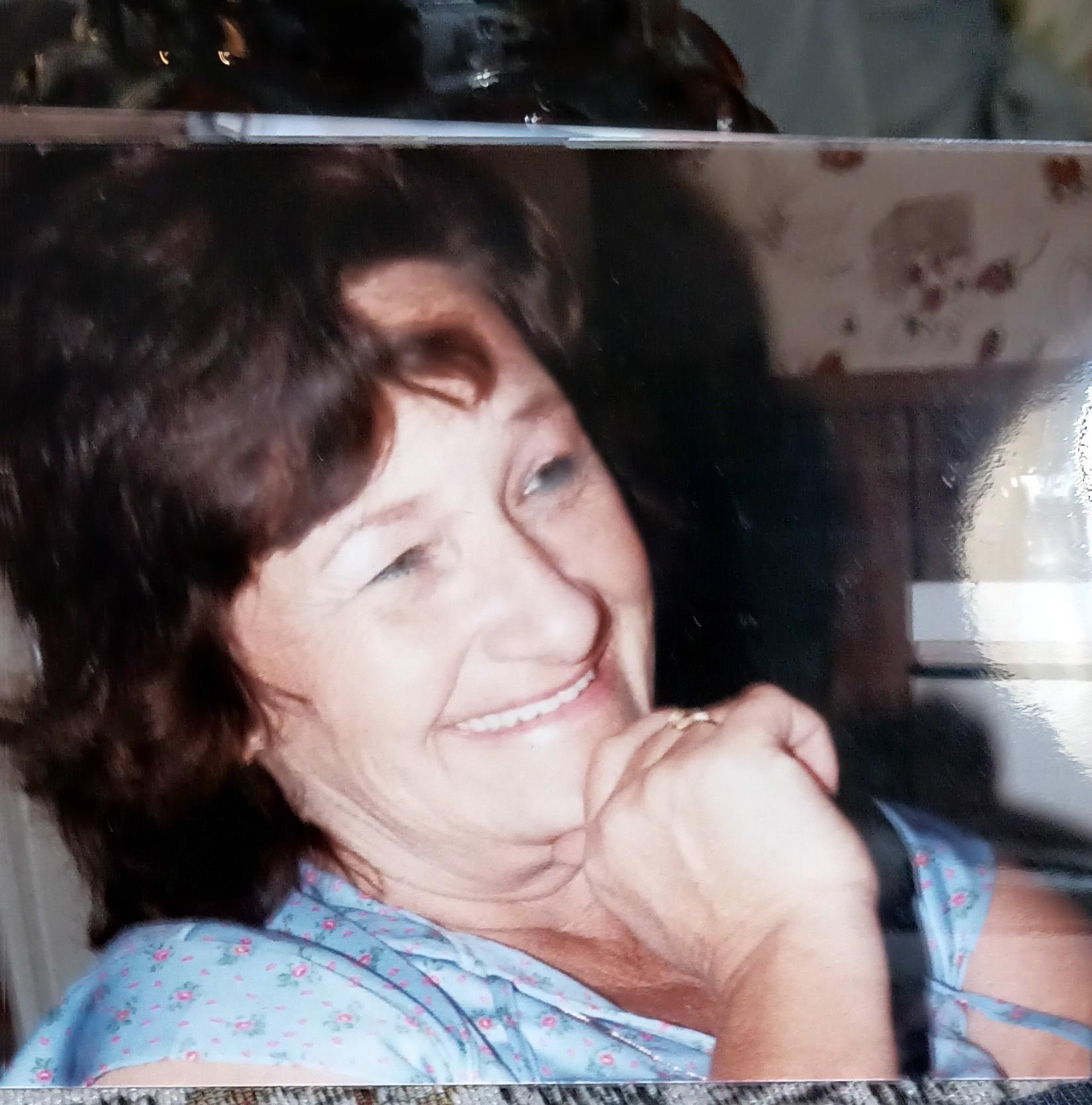 A woman with brown hair smiles while resting her chin on her hand, relaxed at home.