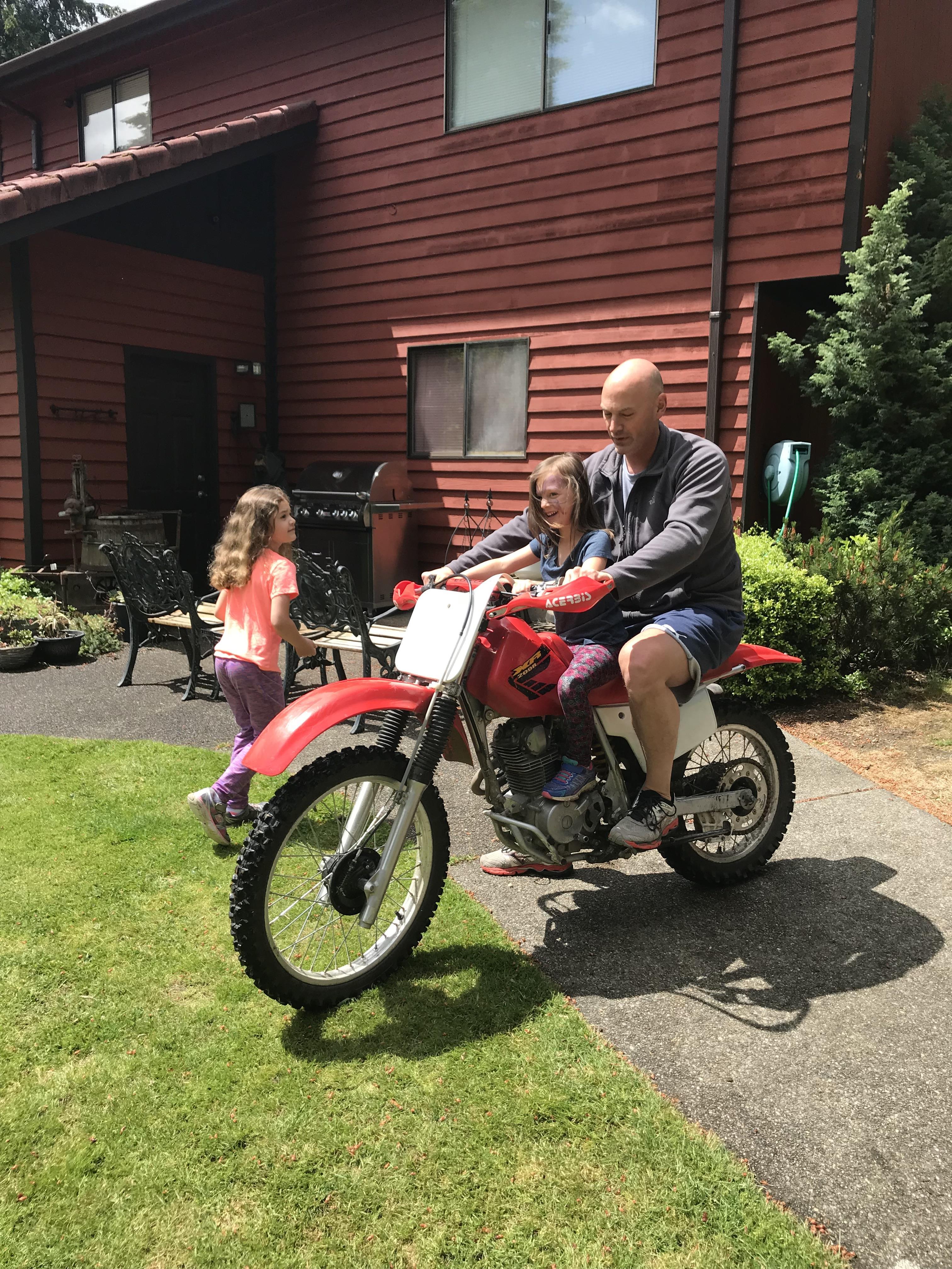 Children laugh joyfully while riding on a dirt bike with their father in a grassy backyard.