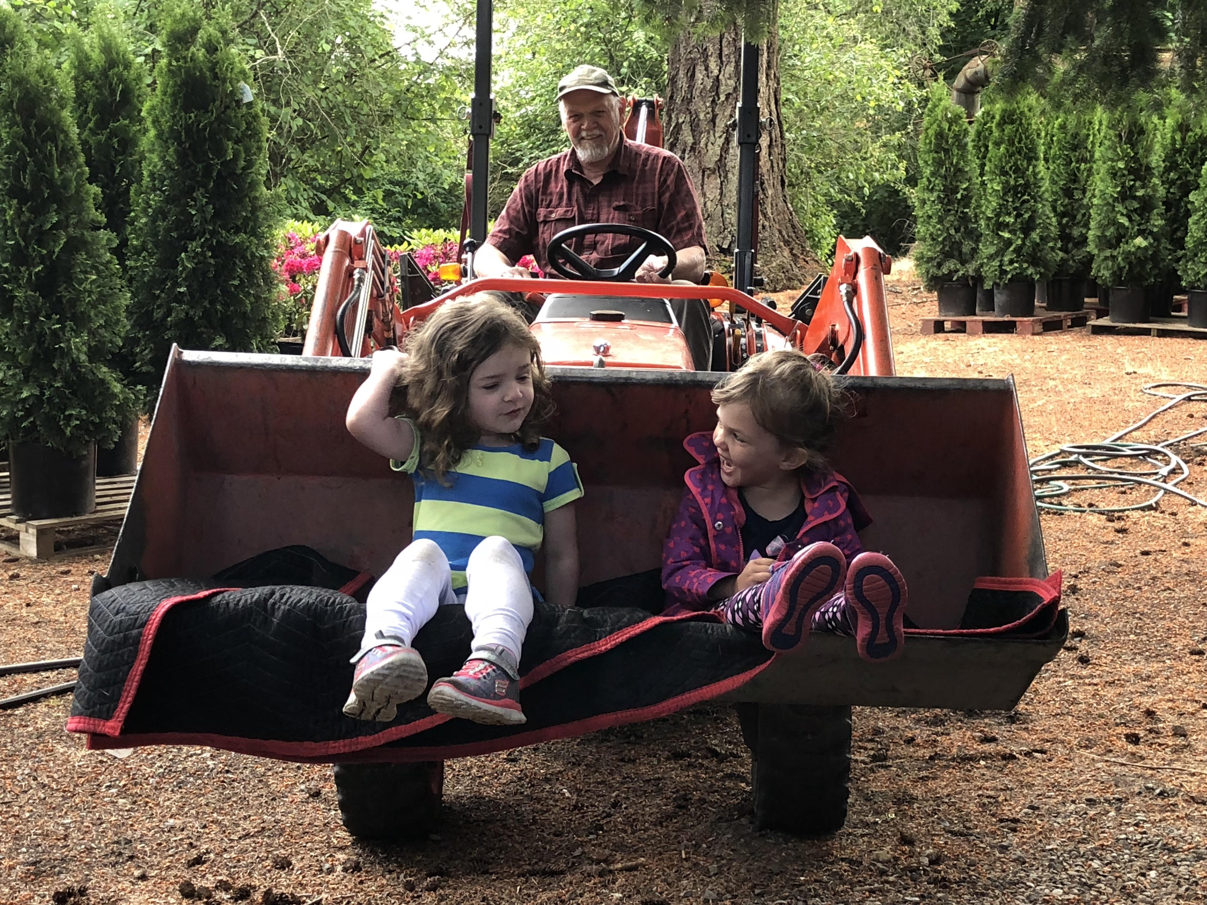 Two girls have fun sitting on a garden tractor while an adult drives through a nursery.