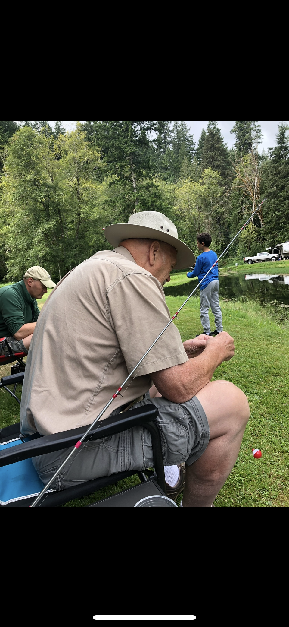 Two men are fishing by a river, while a child plays in the background on a warm afternoon.