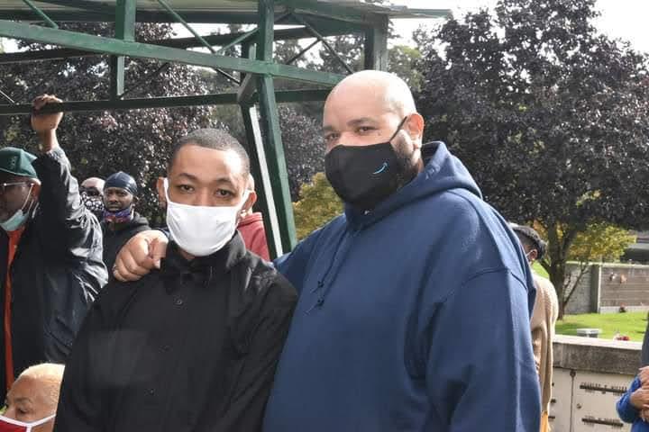 Two men stand together at a community gathering in the park, both wearing masks.