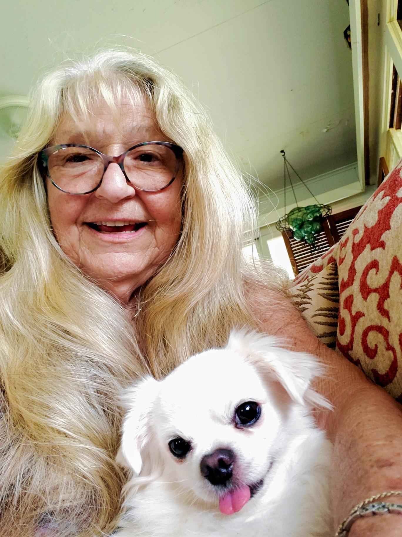 A woman with long hair smiles widely while holding her small white dog on a couch.