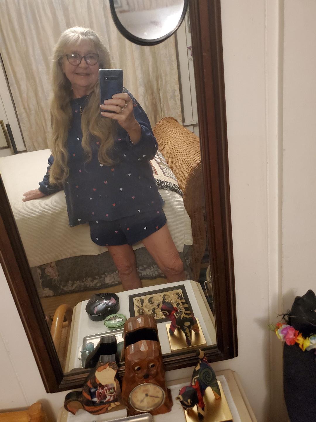 A woman in pajamas stands in a bedroom, smiling while taking a selfie in the mirror.