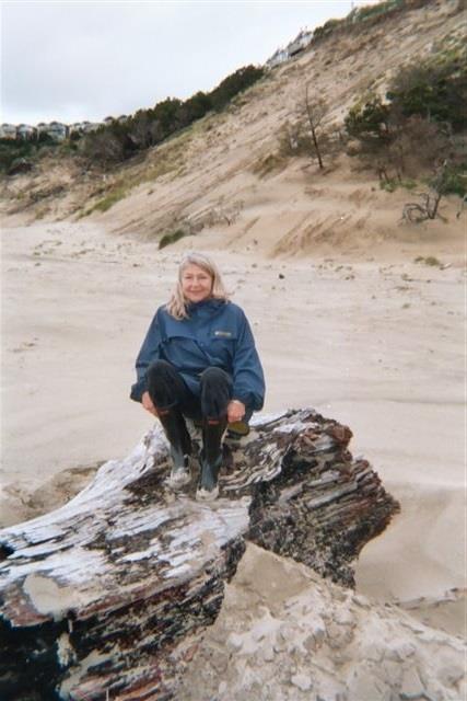 A woman sits on a large log at the beach, smiling against a backdrop of sand dunes and cloudy skies.