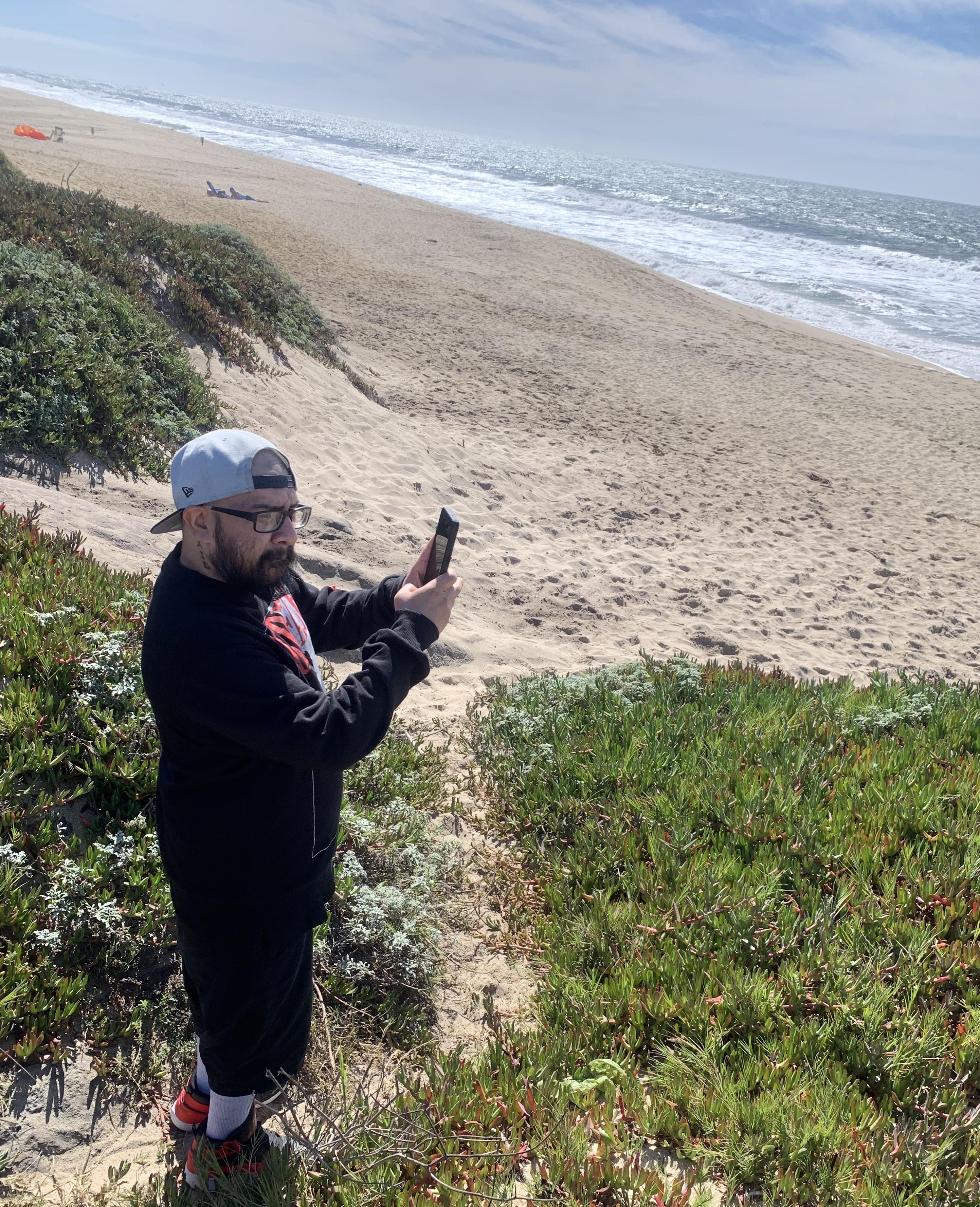 A man stands on the sand with a smartphone, capturing the beach and ocean view on a sunny day.