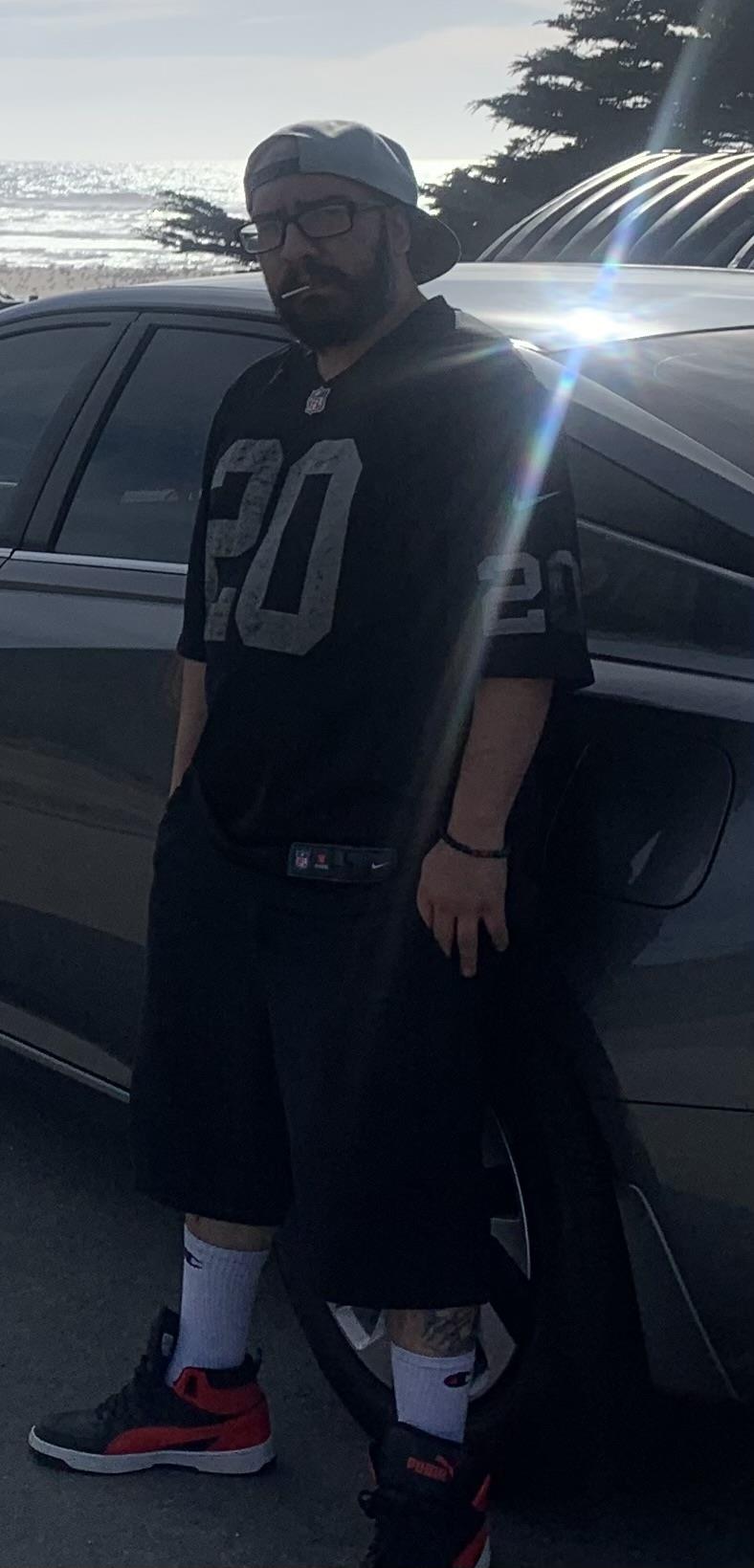 Man stands casually next to a vehicle, dressed in sports gear, at dusk by the coast.