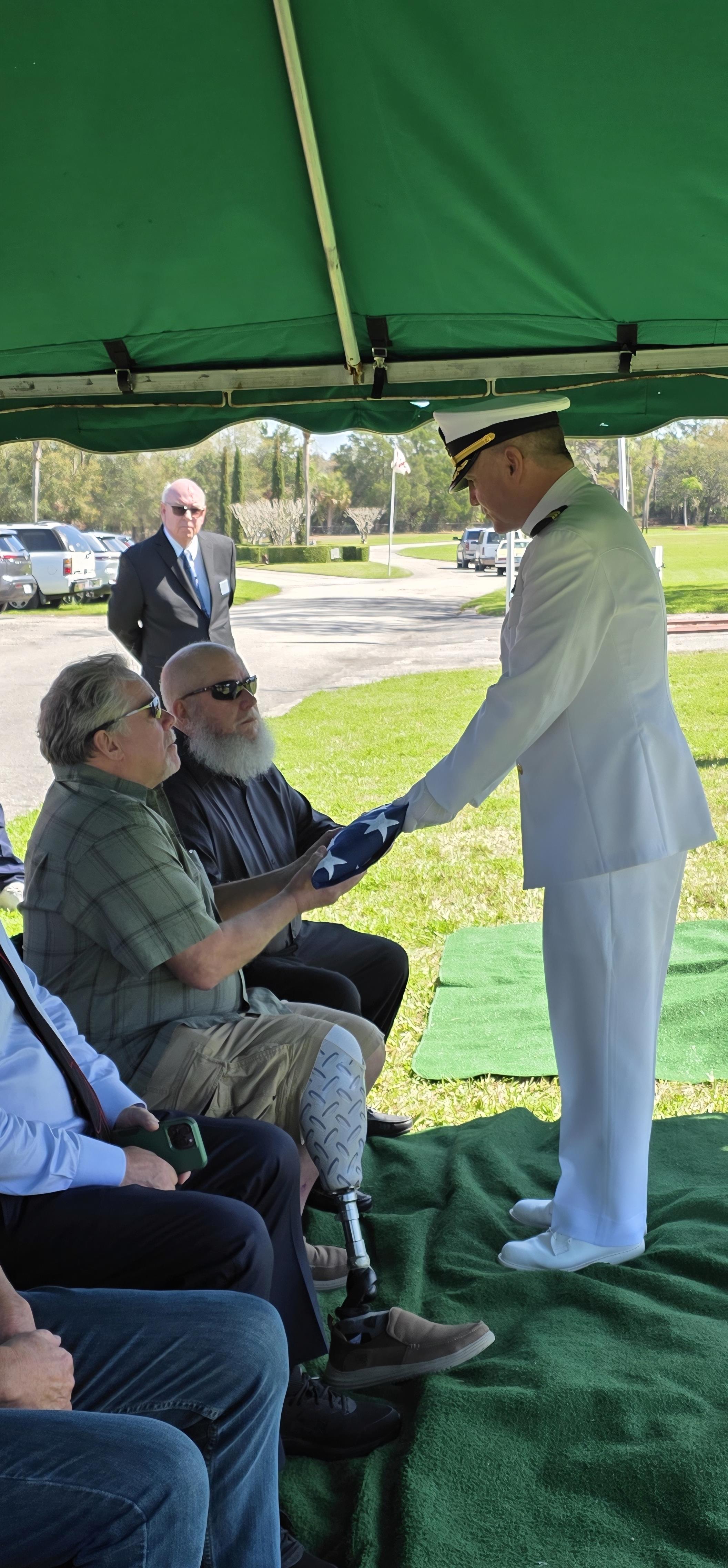 Naval officer presents medals to veterans under a tent at a memorial ceremony in the afternoon.