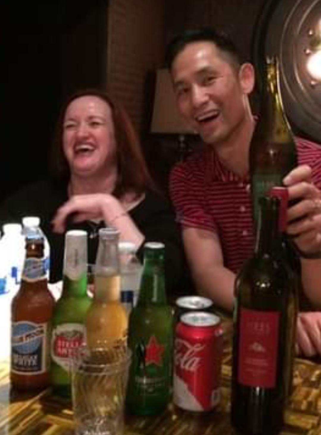 Two friends share a joyful moment, laughing and raising drinks among various beverages on the table.