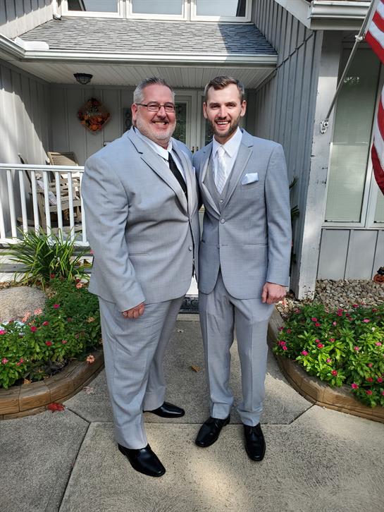 Two men, dressed in matching light grey suits, smile and pose outside a charming home.