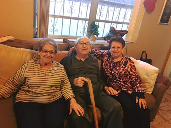 Three older adults enjoy each other's company on a comfortable couch in a homey setting.