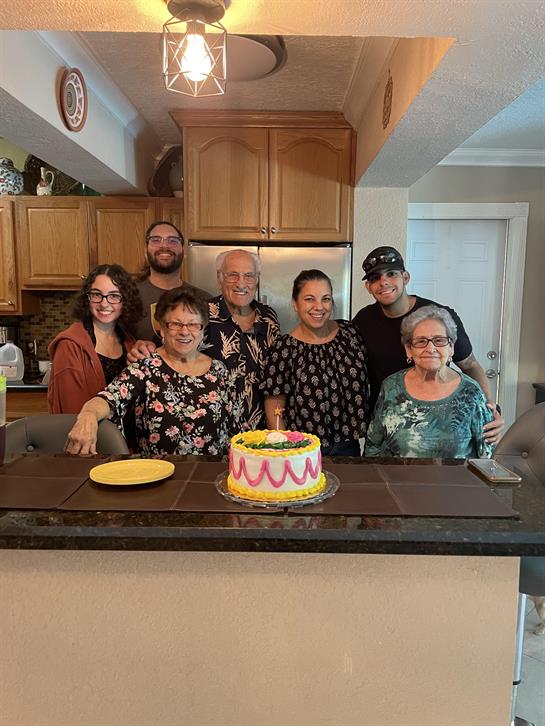 Family members gather around a table with a cake, enjoying a special occasion in a warm kitchen.