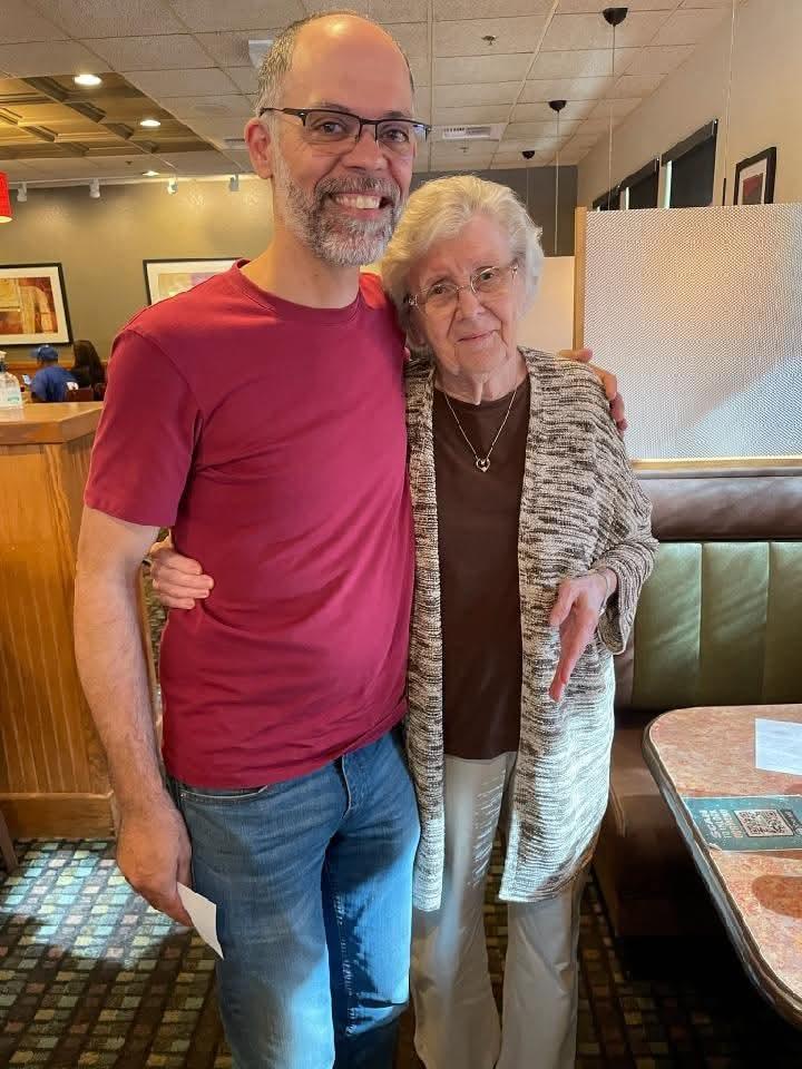 A smiling man embraces an elderly woman at a table in a restaurant, sharing a warm connection.