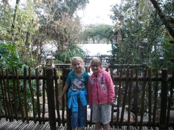 Two girls stand by a wooden fence surrounded by greenery, smiling while enjoying the outdoors.
