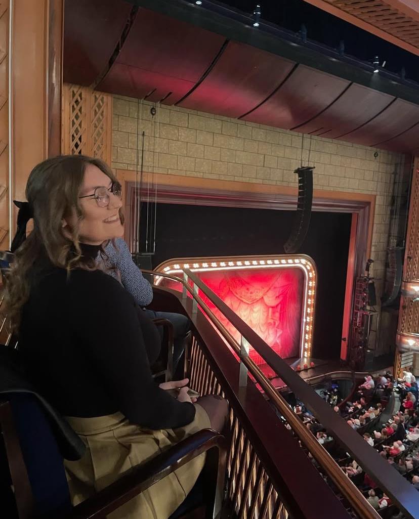 A young woman relaxes in a balcony seat, watching a live performance in a grand theater.
