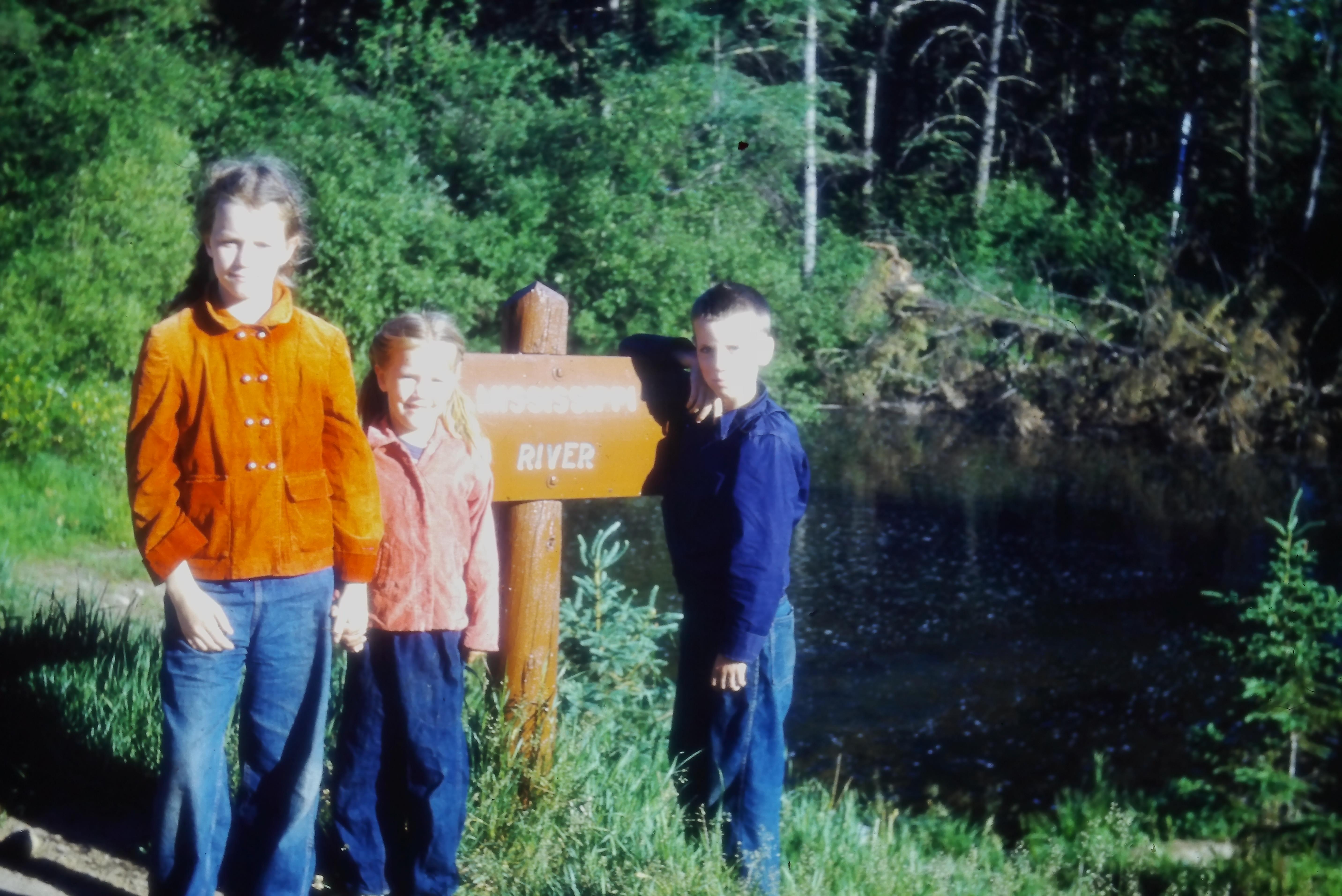 Three children pose next to a sign by a tranquil lake surrounded by lush trees.