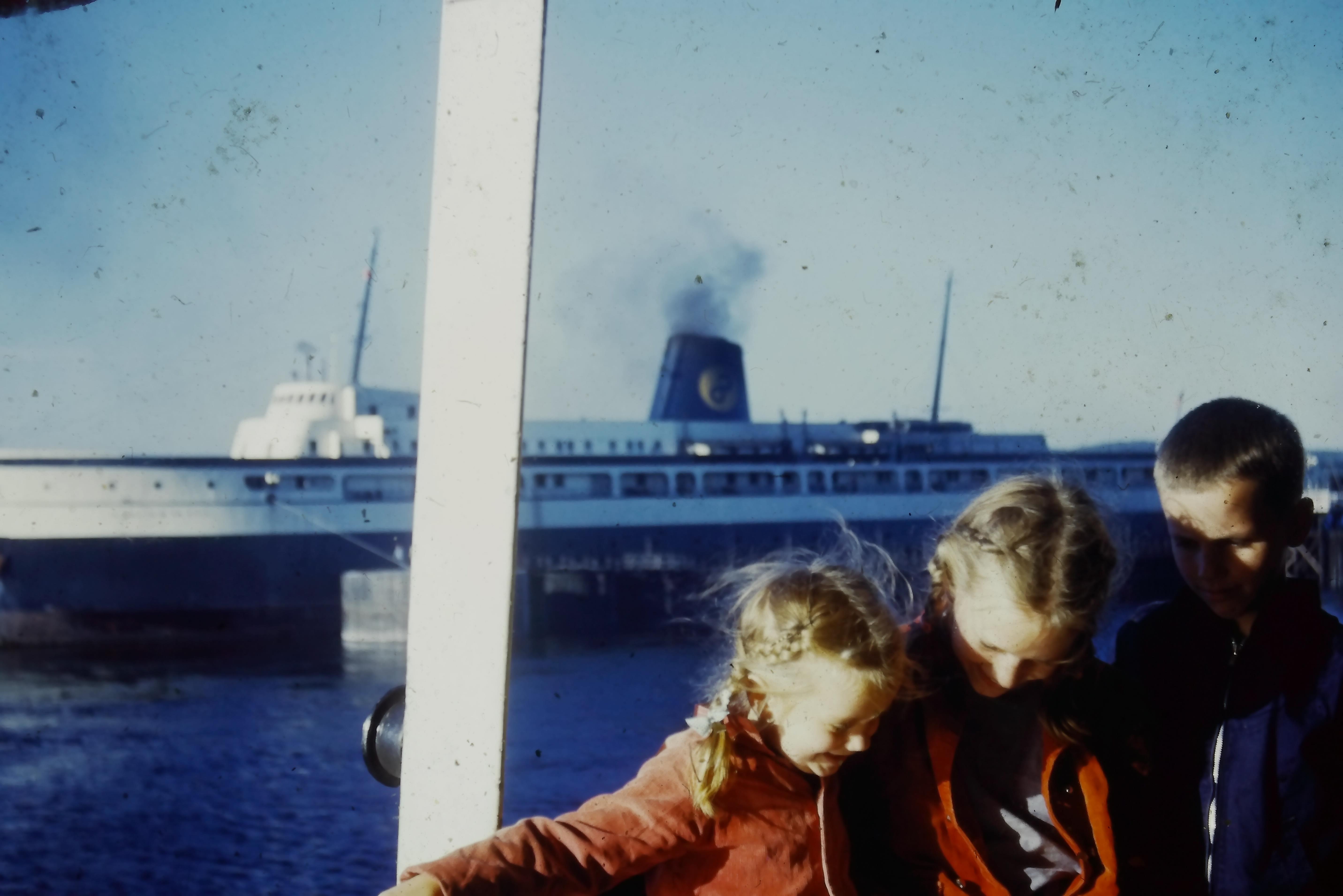 Three children gather on a dock, smiling and leaning against each other as a ship passes by.