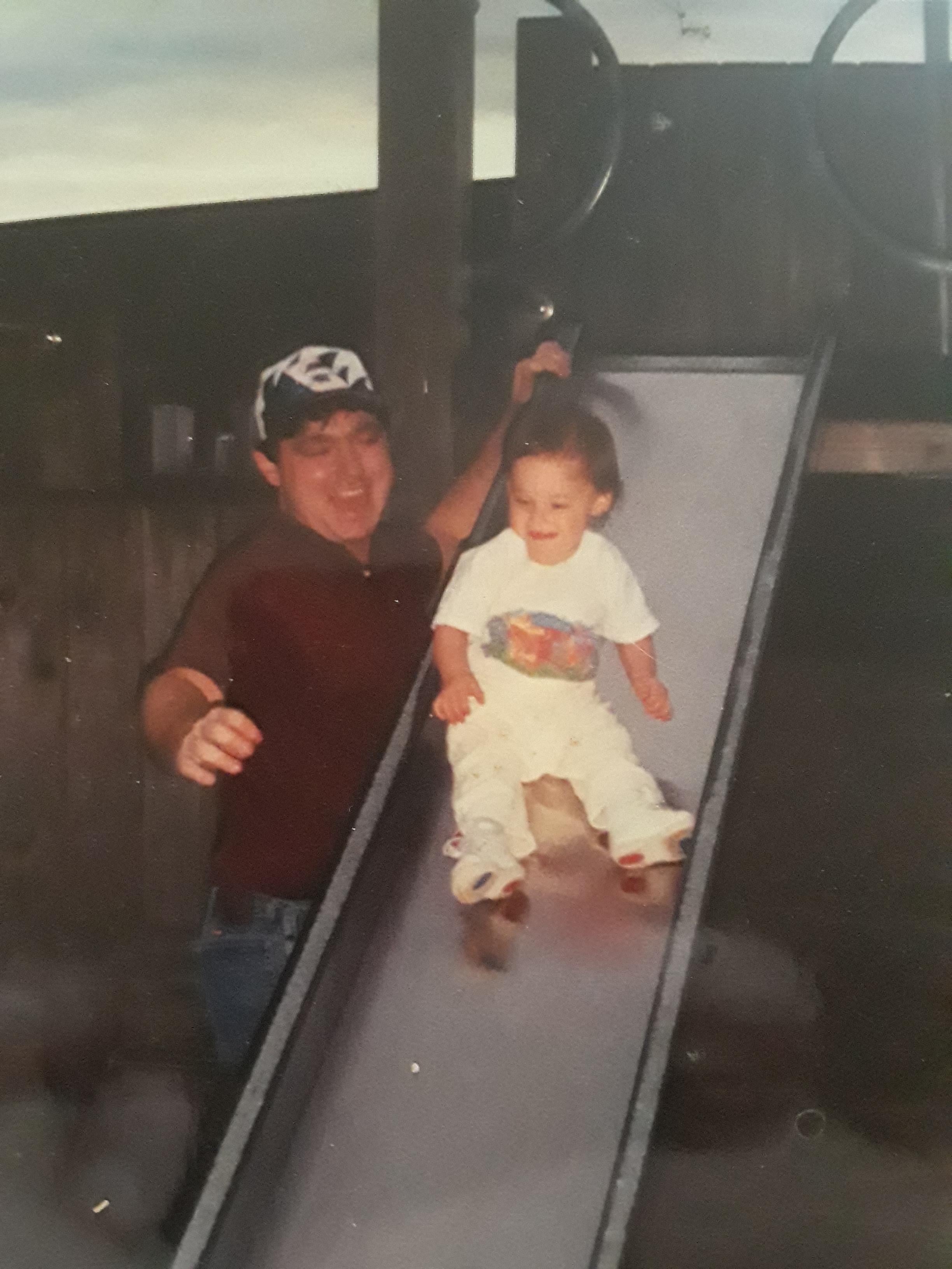 A man helps a young child slide down a playground slide on a cloudy day, filled with laughter.