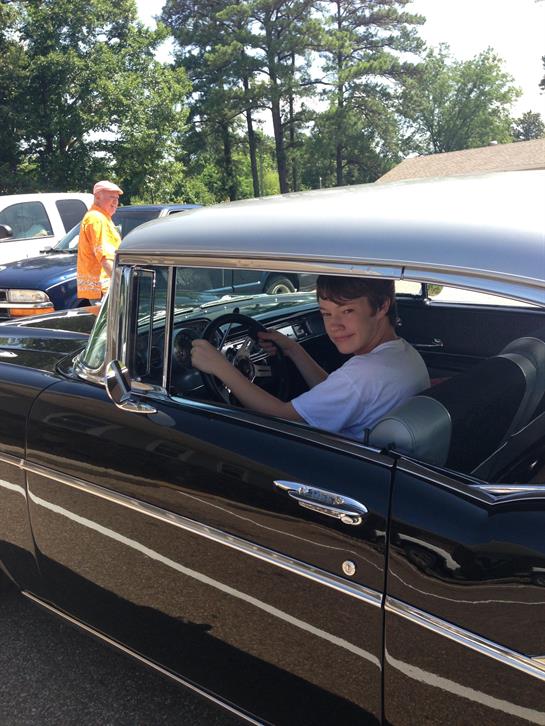 A boy smiles while seated behind the wheel of a vintage car during a summer gathering.