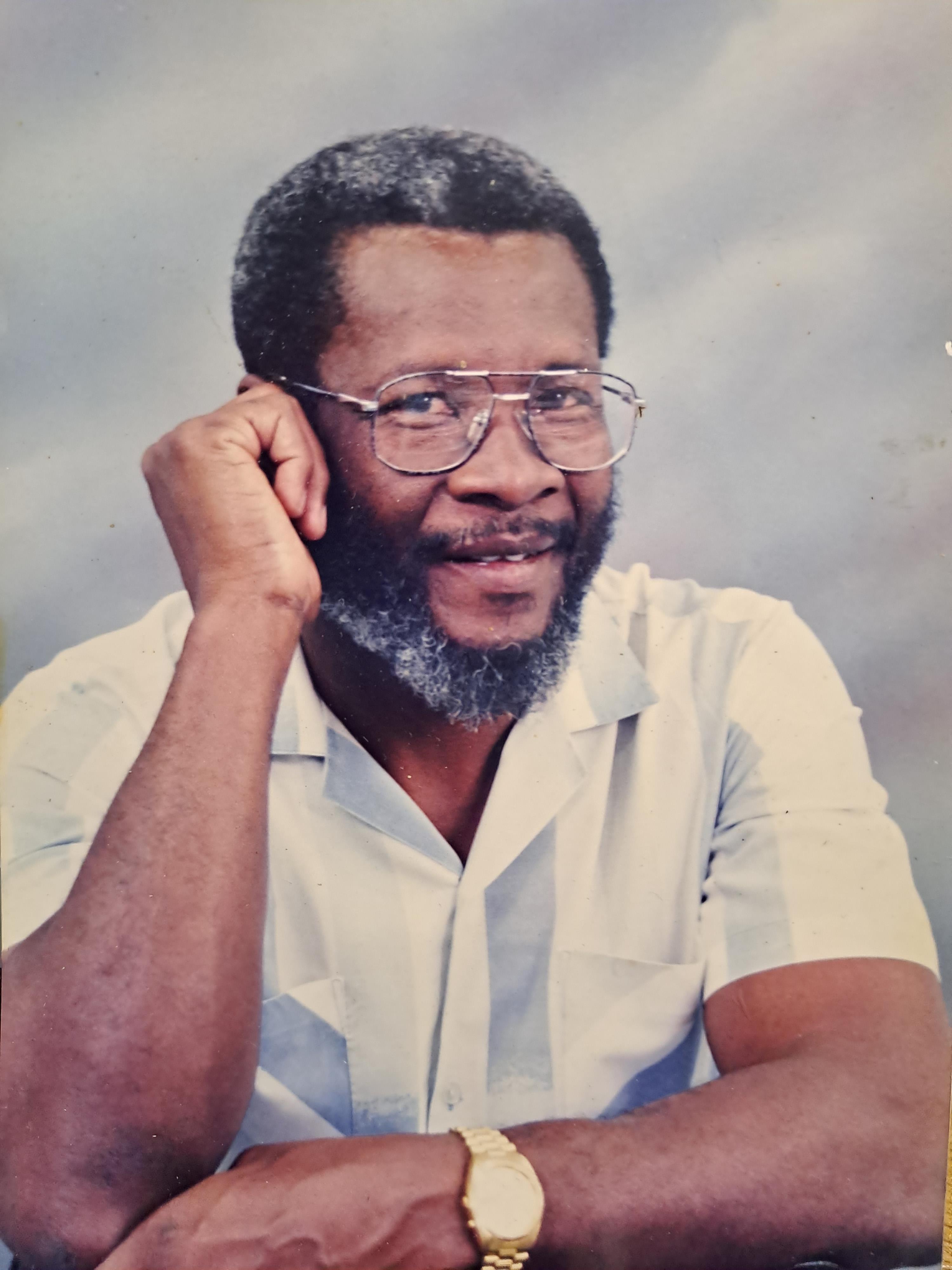 A man with glasses smiles broadly while resting his chin on his hand, wearing a light shirt.