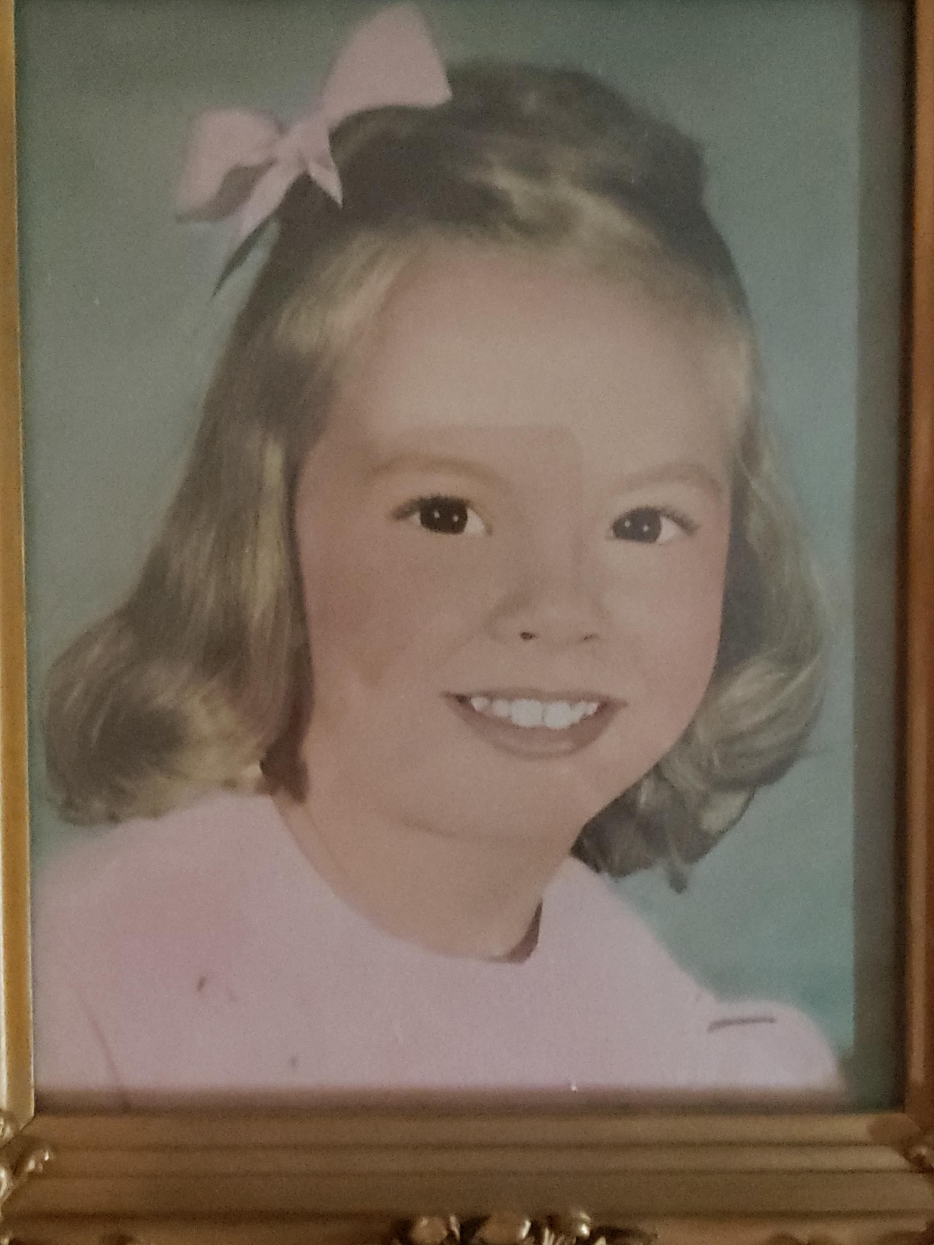 A cheerful young girl smiles widely, dressed in a light pink outfit with a bow in her hair.
