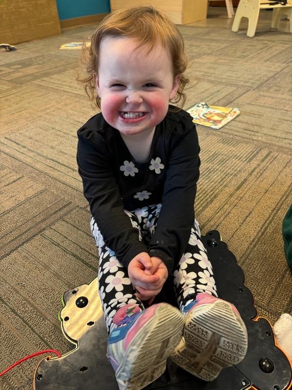 A cheerful young girl enjoys her time sitting on a colorful mat in a playroom, smiling widely.
