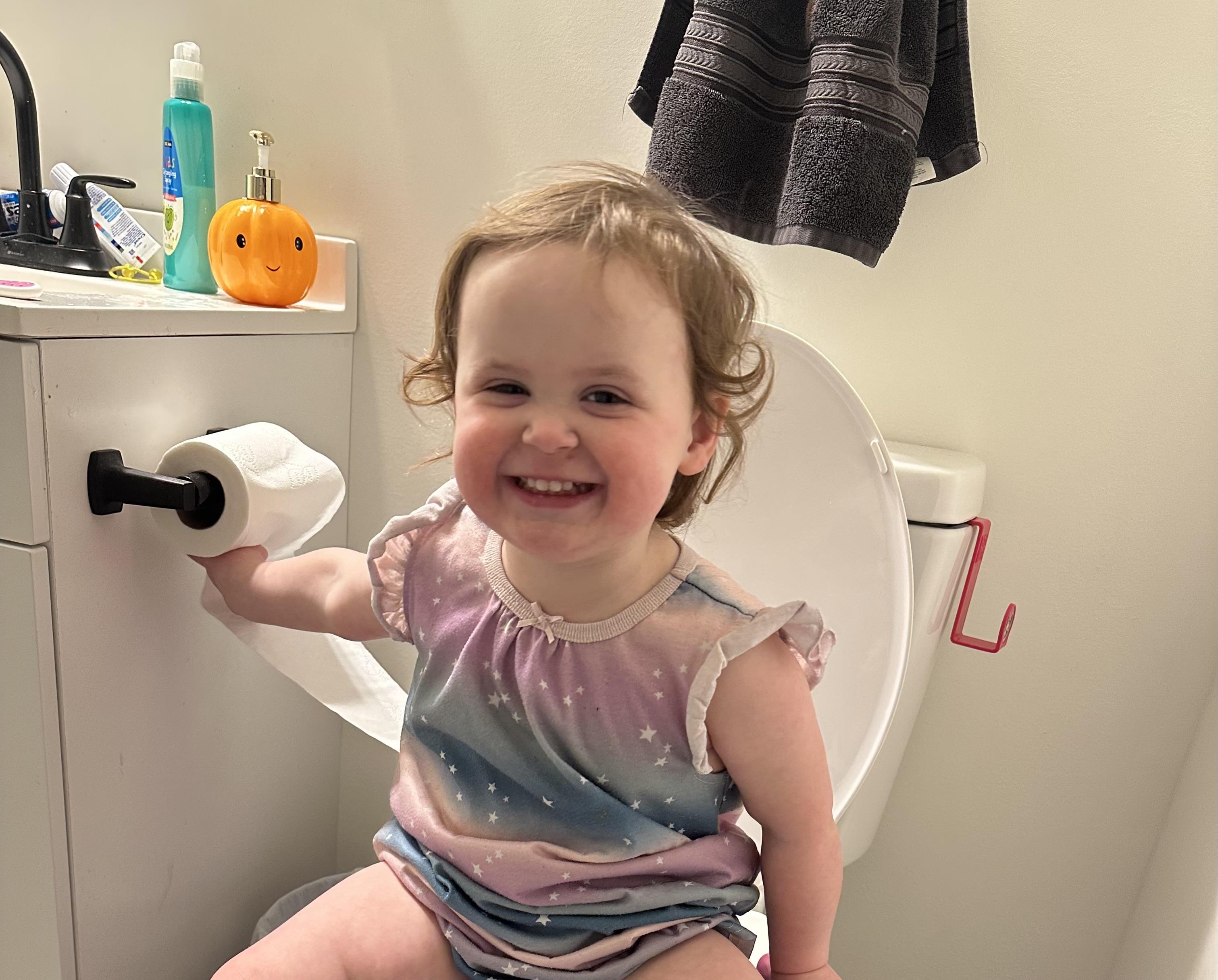 Toddler smiles joyfully in bathroom as she engages with toilet and holds a roll of paper.
