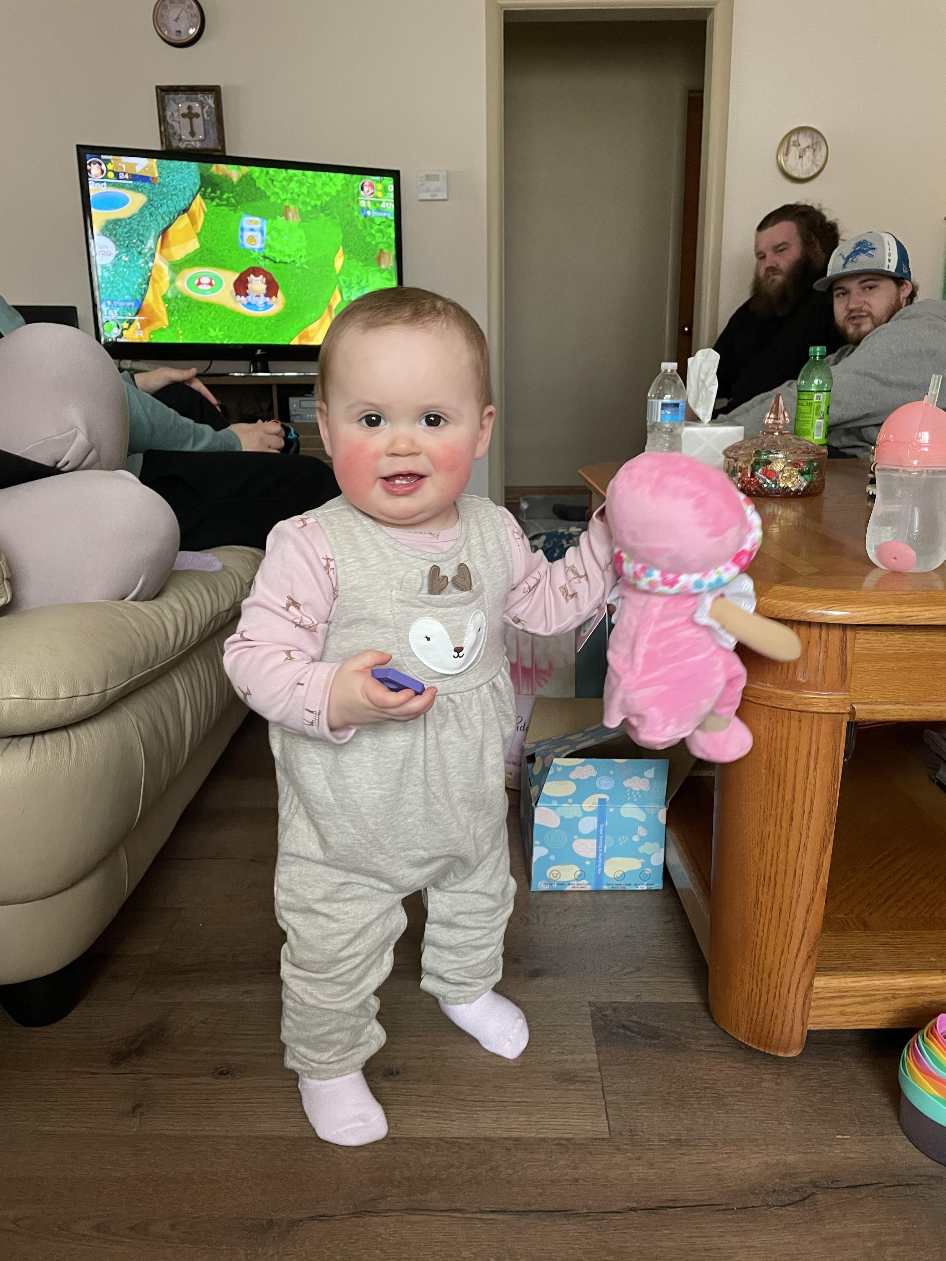 A baby with a cheerful expression stands holding a pink doll in a cozy living room.