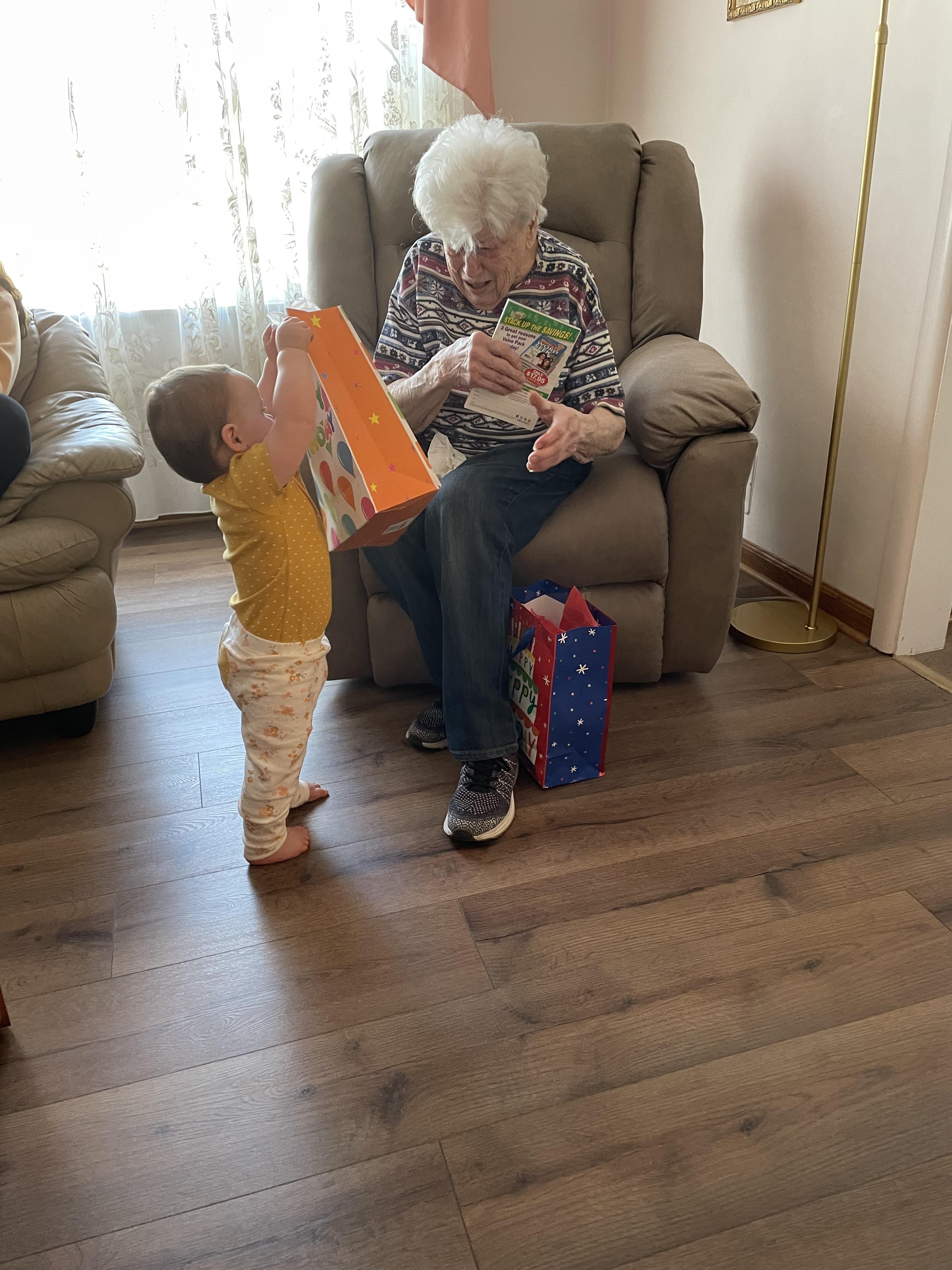 A toddler excitedly hands a gift to an elderly woman while seated in a comfortable living room.