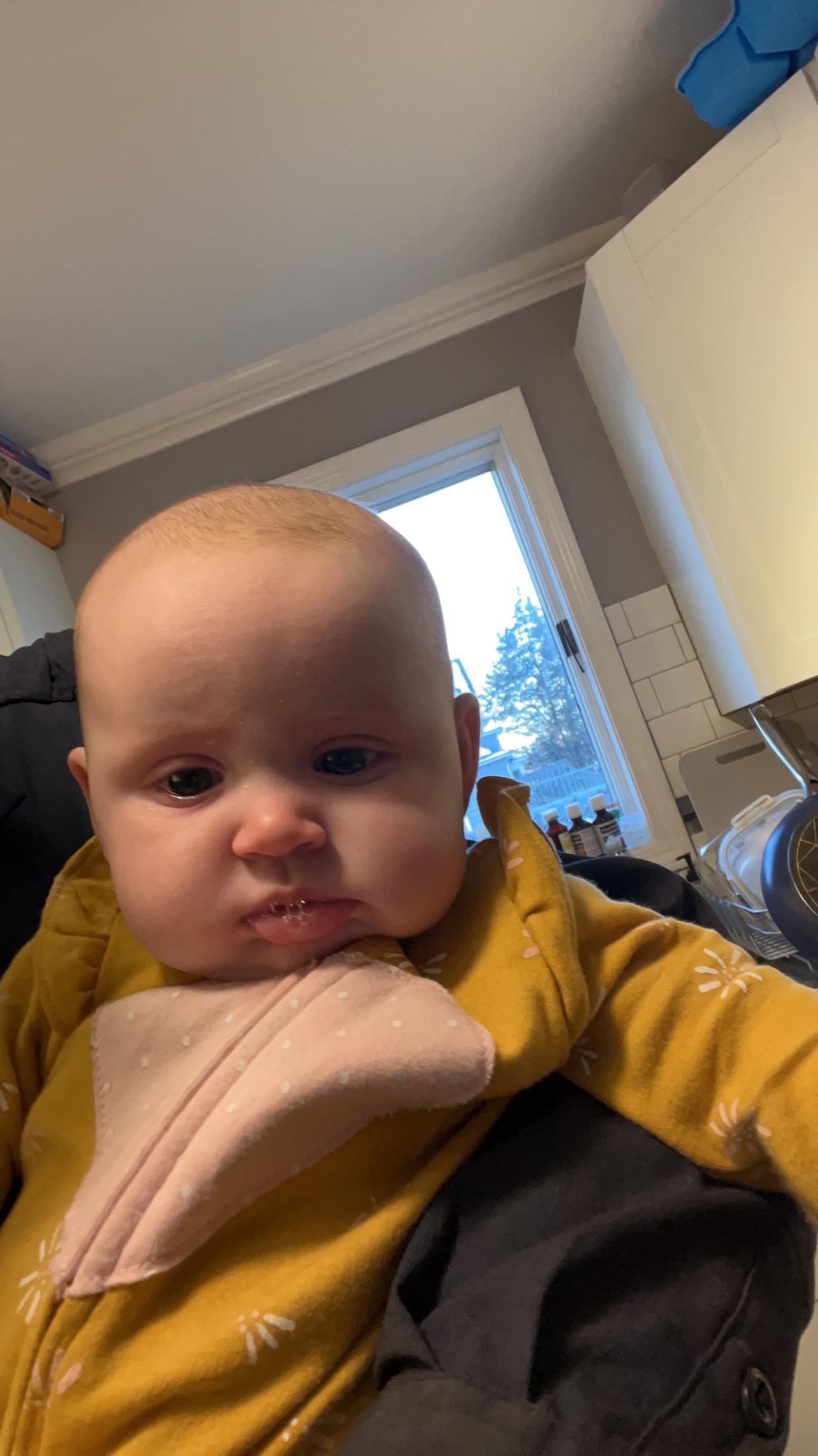 A baby in a cozy outfit is being held in a warm kitchen with snowy view outside.