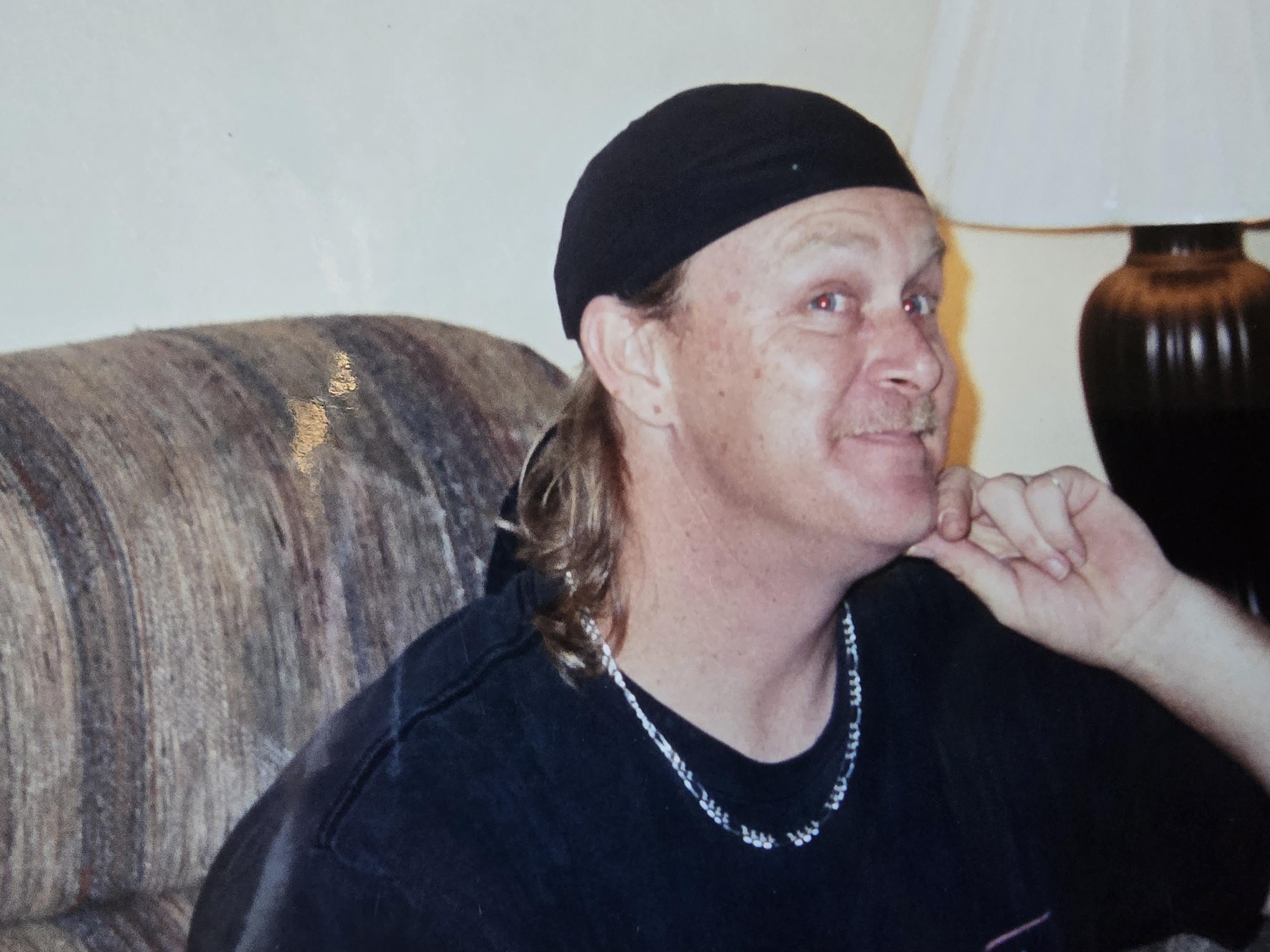 A man wearing a black hat and necklace sits comfortably on a couch, smiling and relaxed.