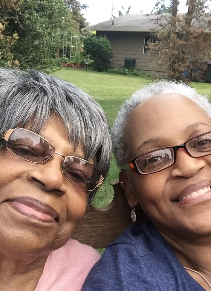 Two joyful women share smiles while sitting on a bench in a green yard under sunlight.