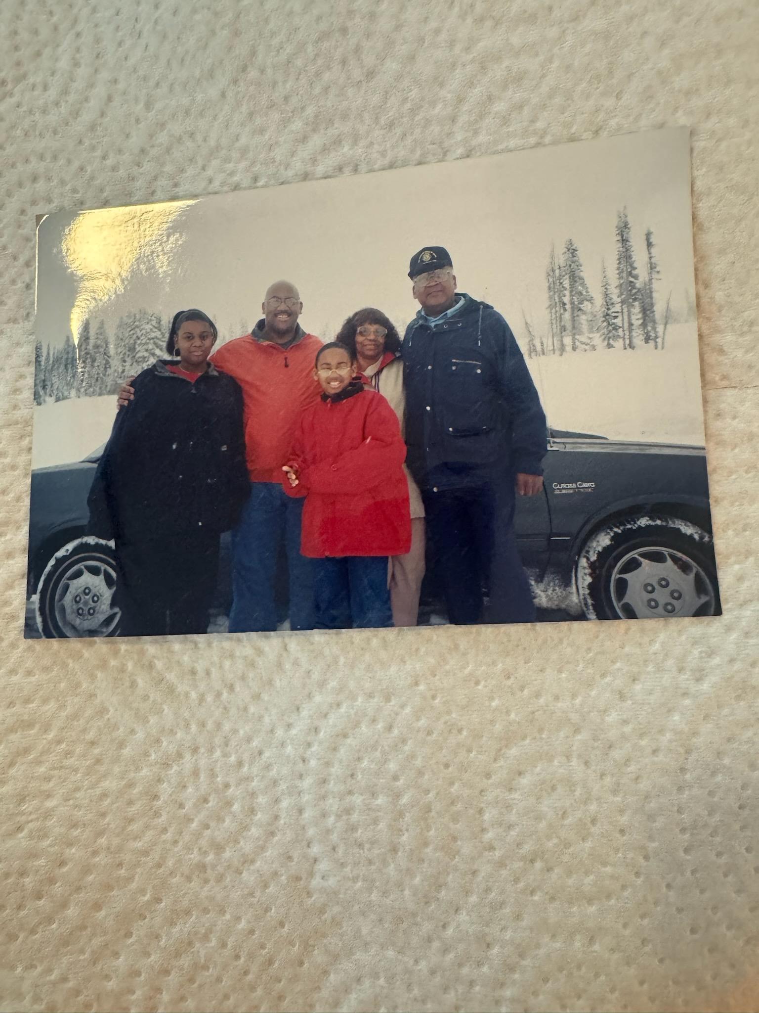 Four friends posing together in winter clothing next to a parked car in a snowy area.