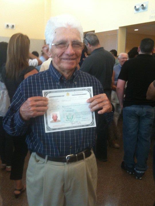 An elderly man displays a certificate surrounded by a bustling crowd in a community center.