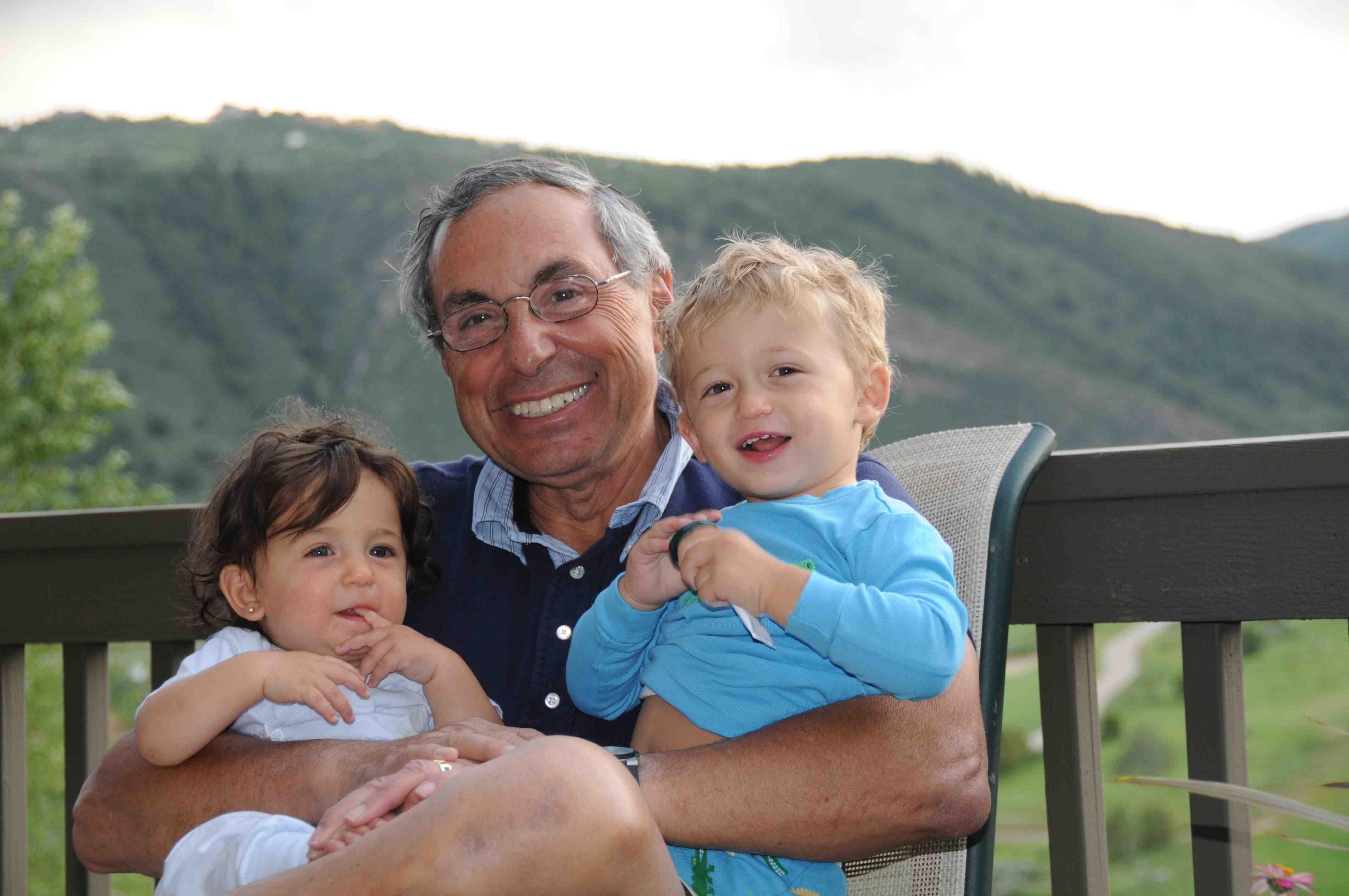 Grandfather smiles warmly with two toddlers, framed by green mountains and a clear sky.