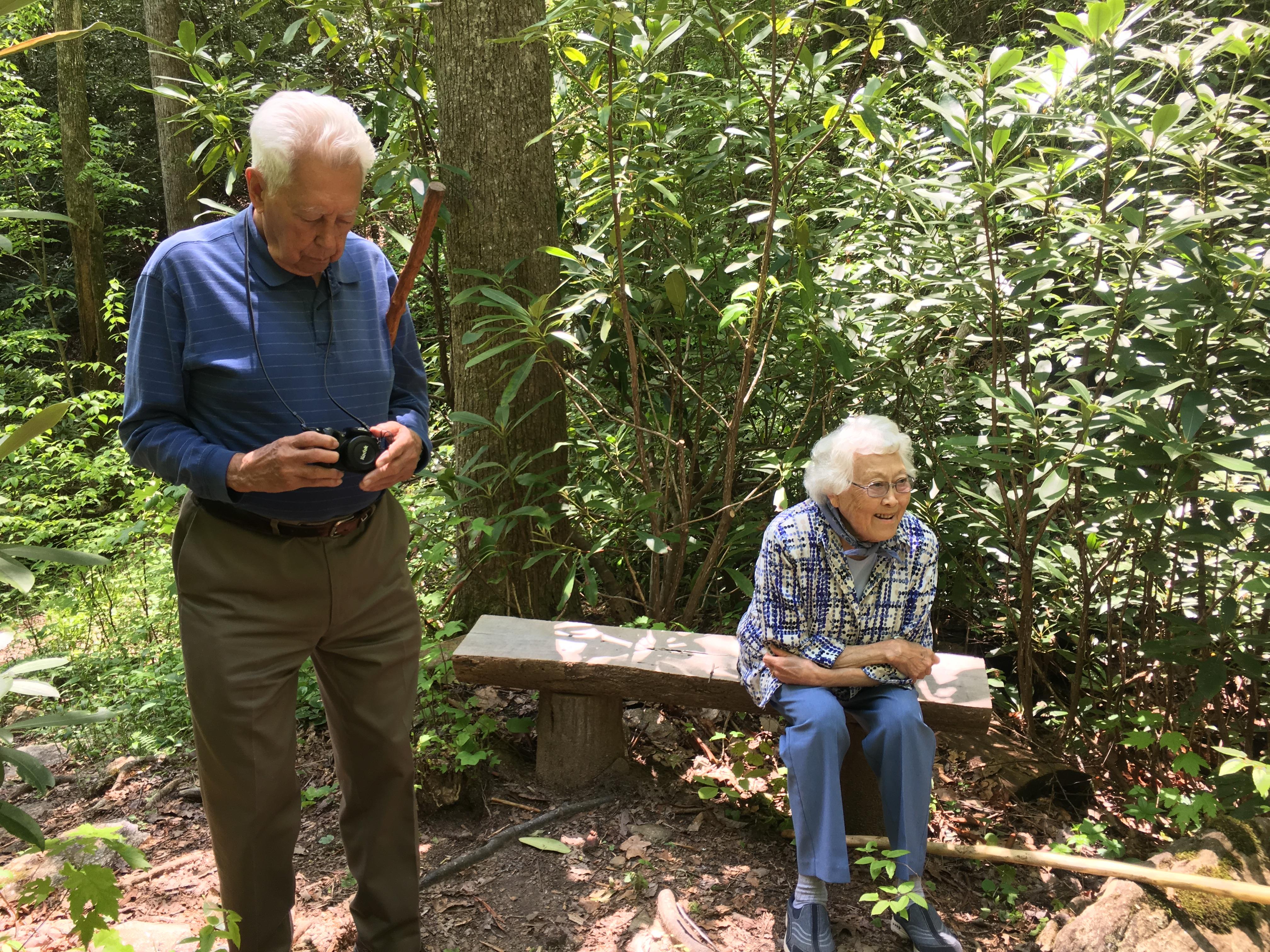 A senior man takes photos while a senior woman sits peacefully on a bench surrounded by greenery.
