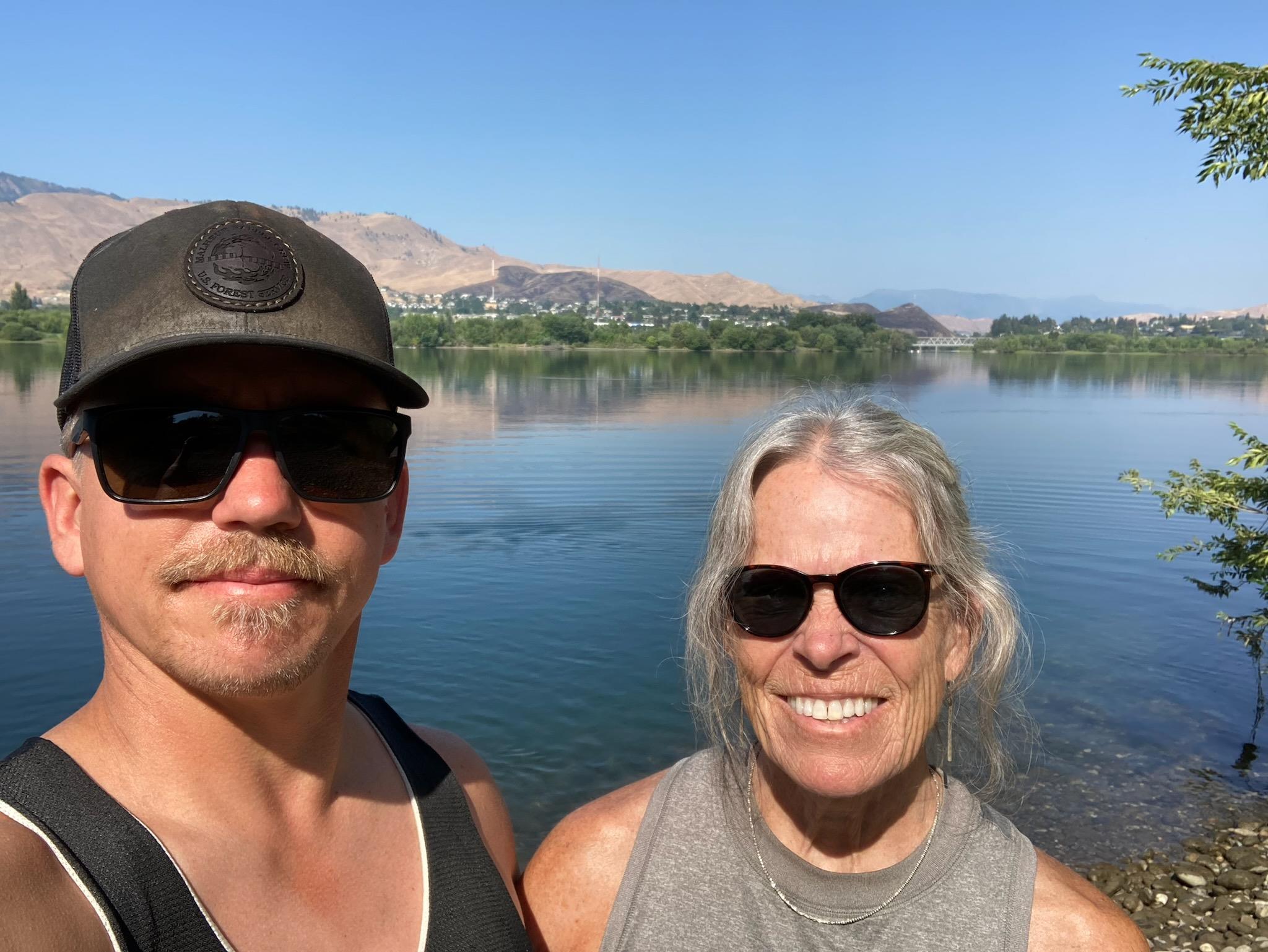 Two friends stand by the lake, smiling and enjoying the warm weather during daylight hours.