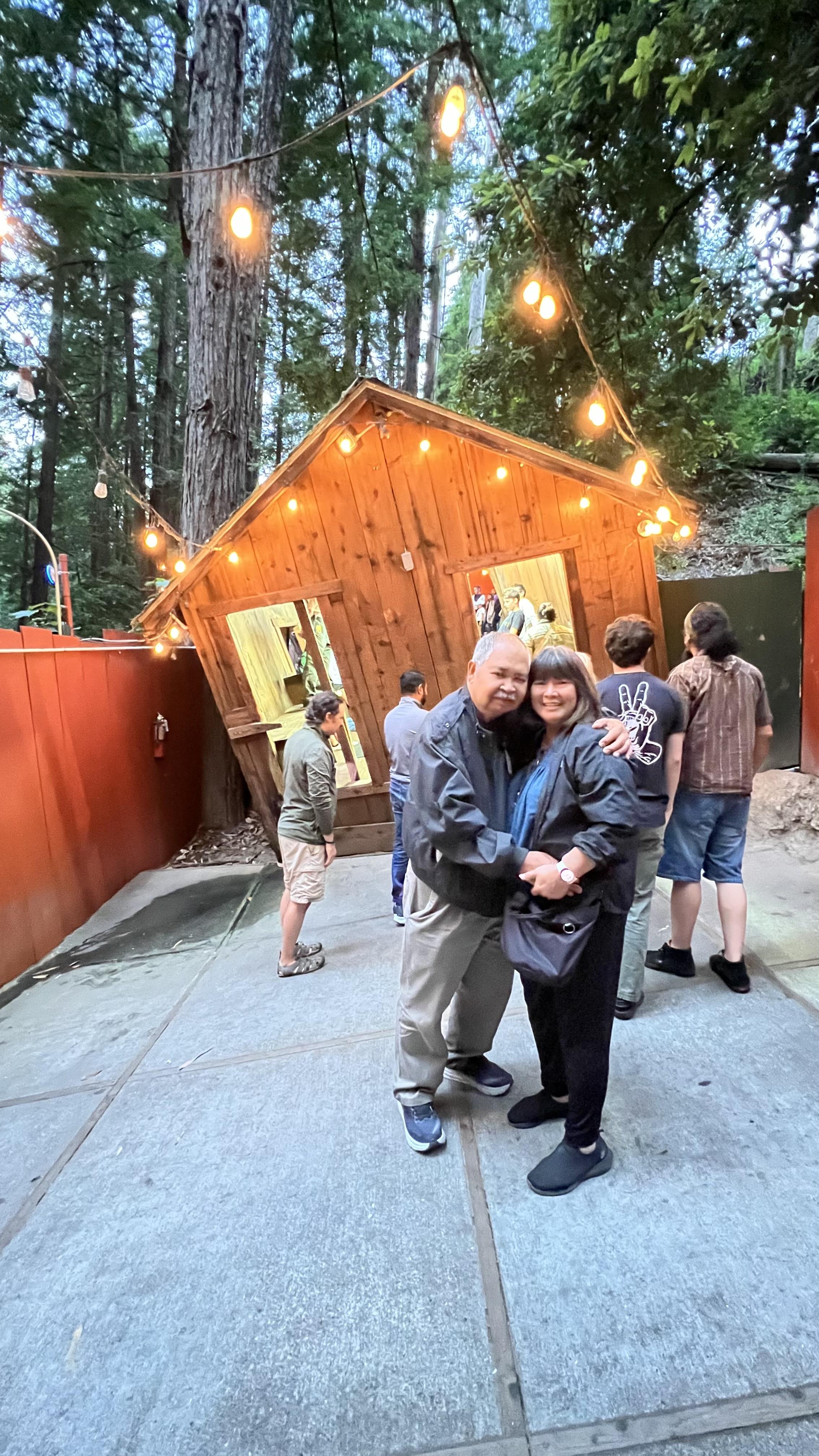 An affectionate couple stands smiling in front of a unique tilted building, surrounded by people.