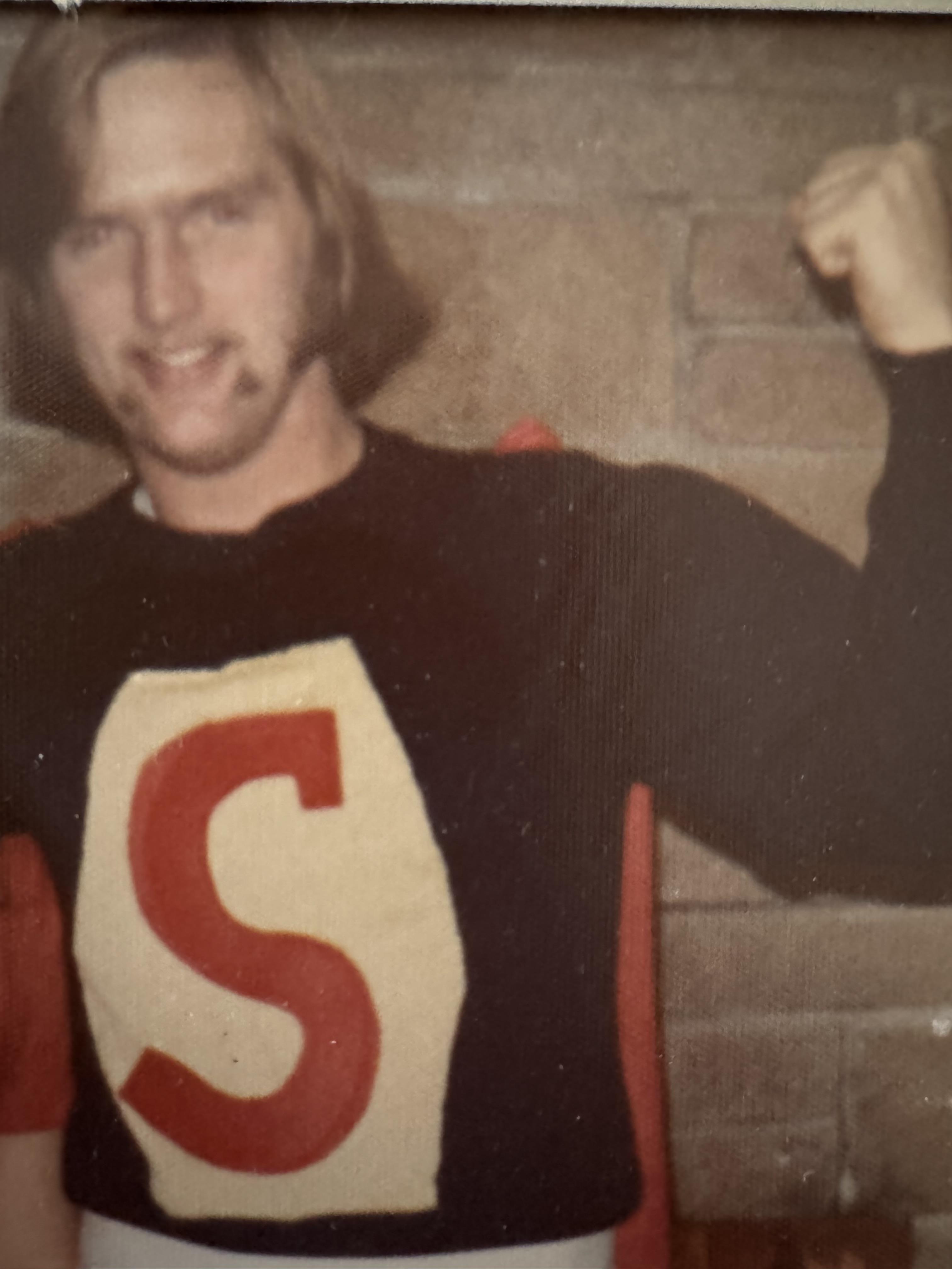 Man in a superhero costume shows off muscle while posing against a textured brick wall.