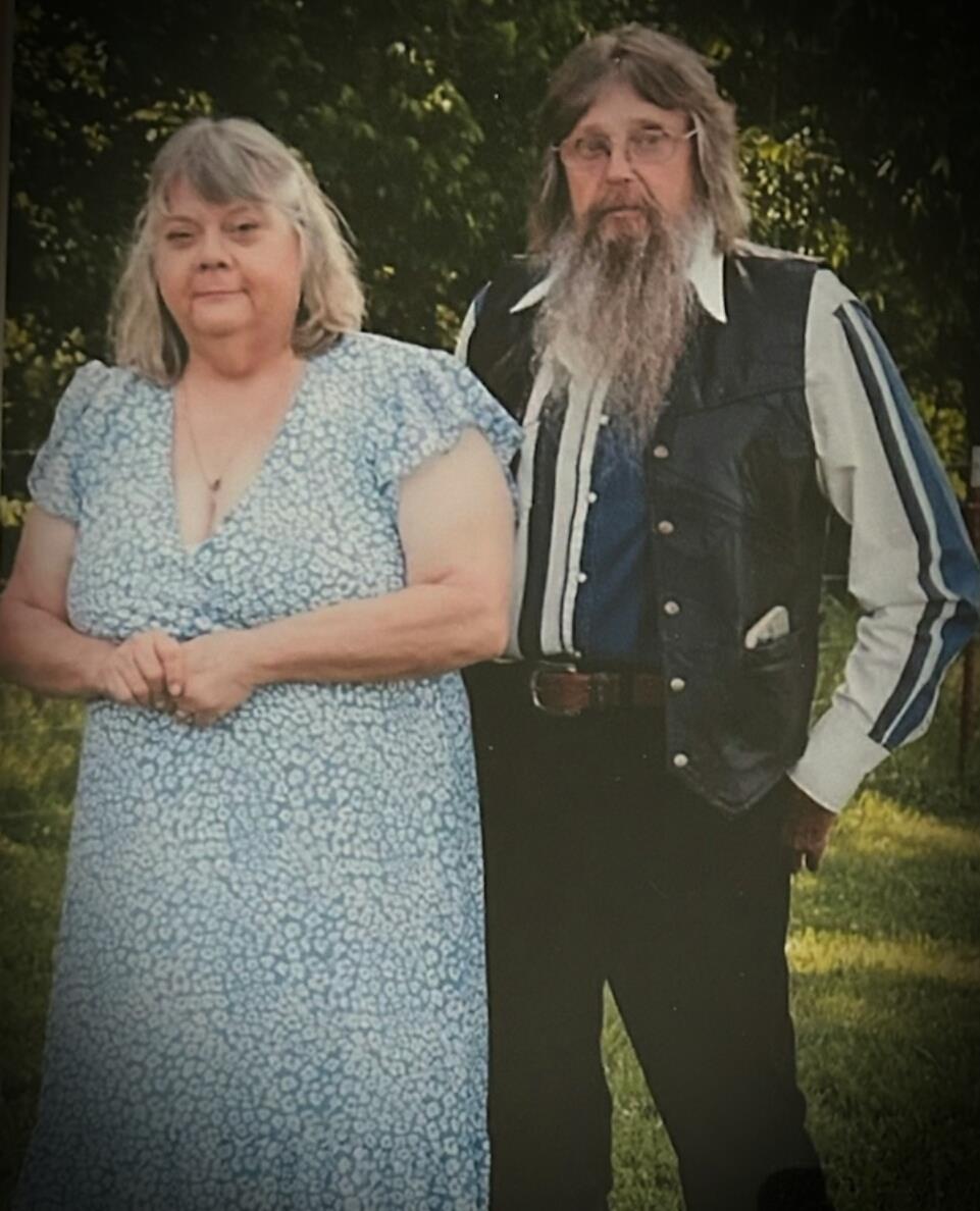A couple stands together, smiling casually in a green outdoor setting under natural light.
