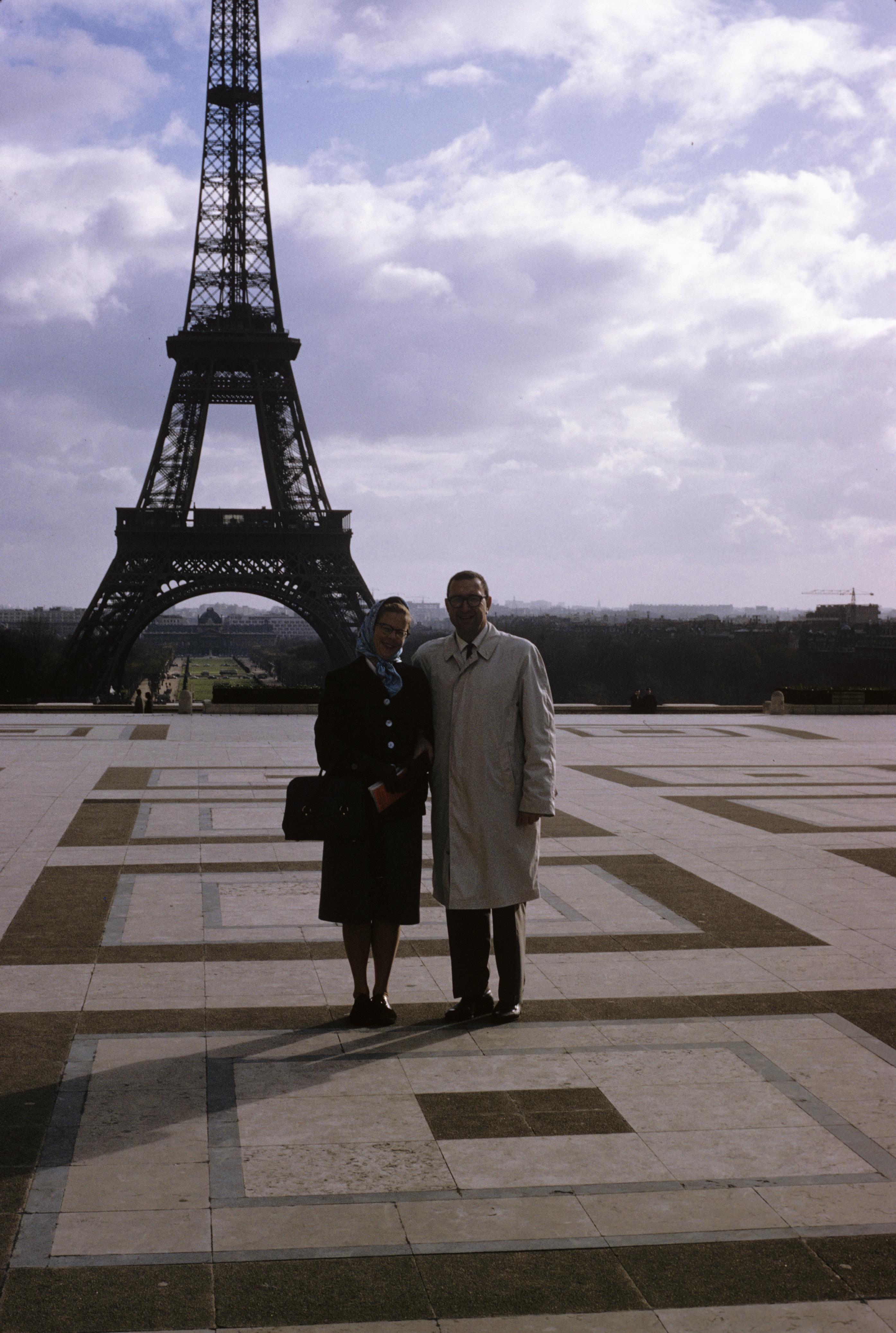 A couple stands together near the Eiffel Tower, dressed warmly on a cloudy day in Paris.