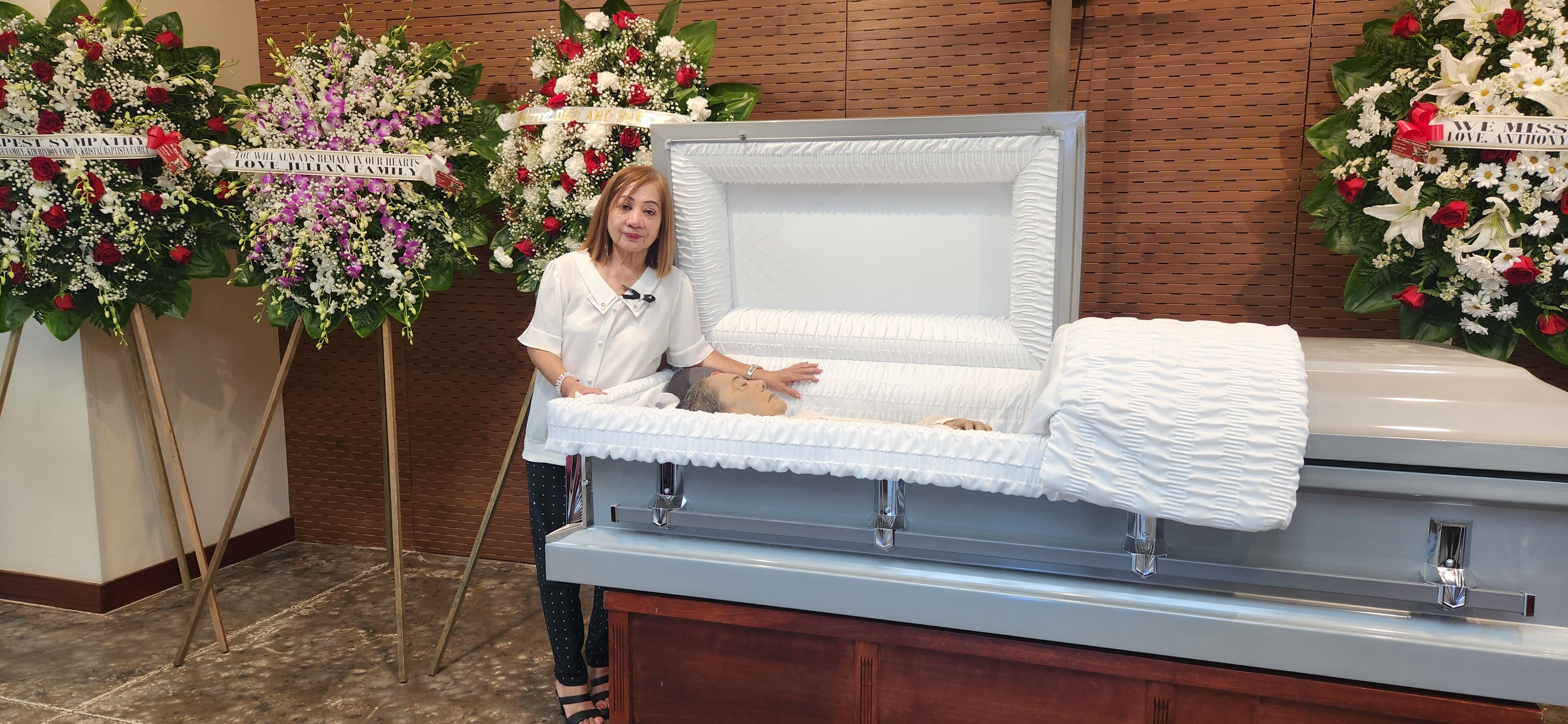Family member stands beside an open casket adorned with flowers in a funeral setting.