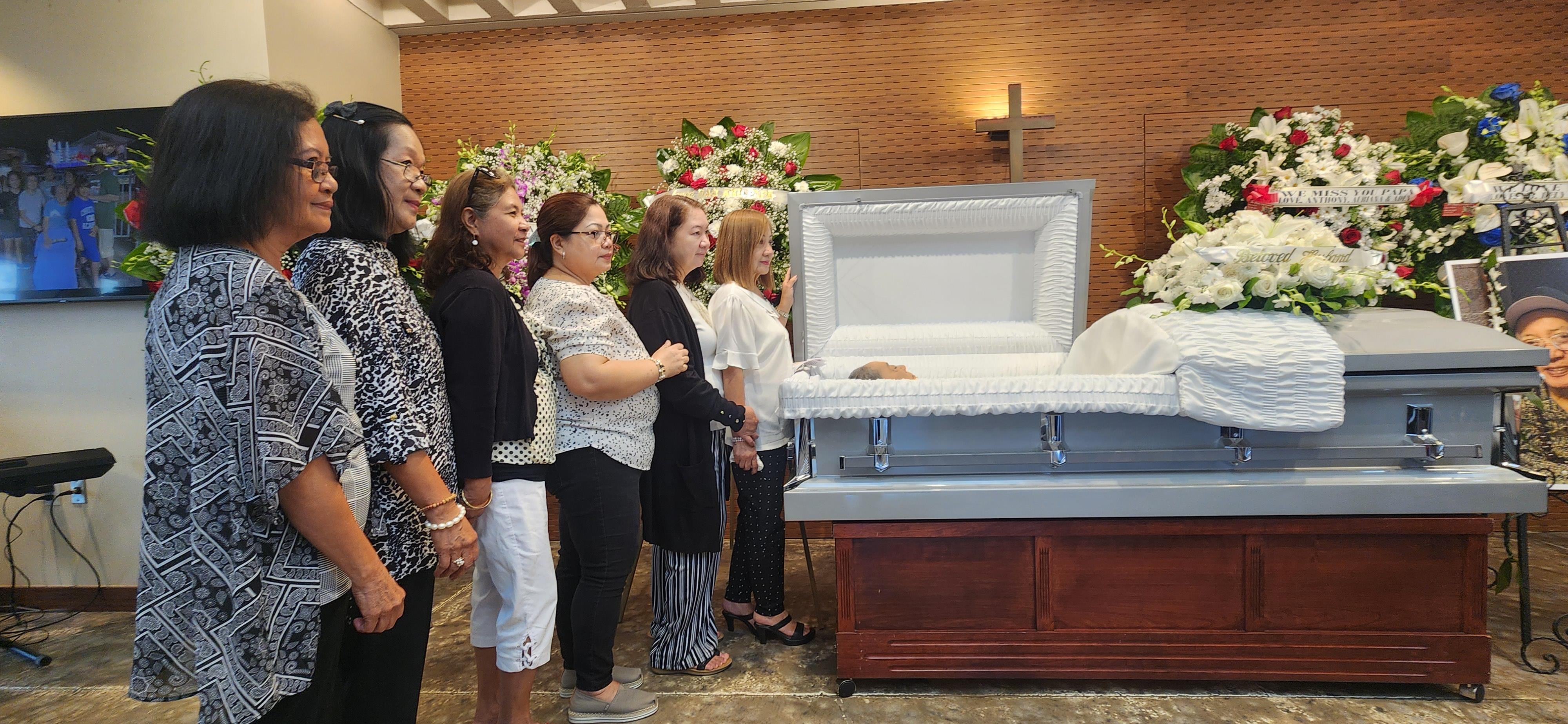 Mourners gather in a quiet space, standing beside a casket adorned with flowers during a memorial.