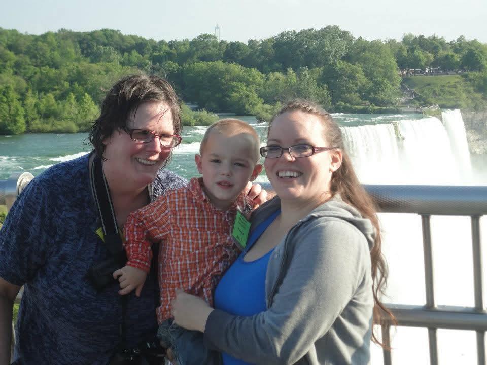 Three people smile joyfully together, with Niagara Falls cascading behind them on a bright day.