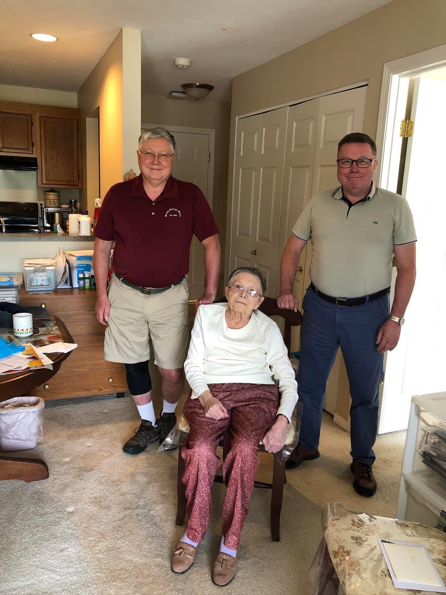 Two men visit their elderly relative in her cozy home, sharing a moment together.