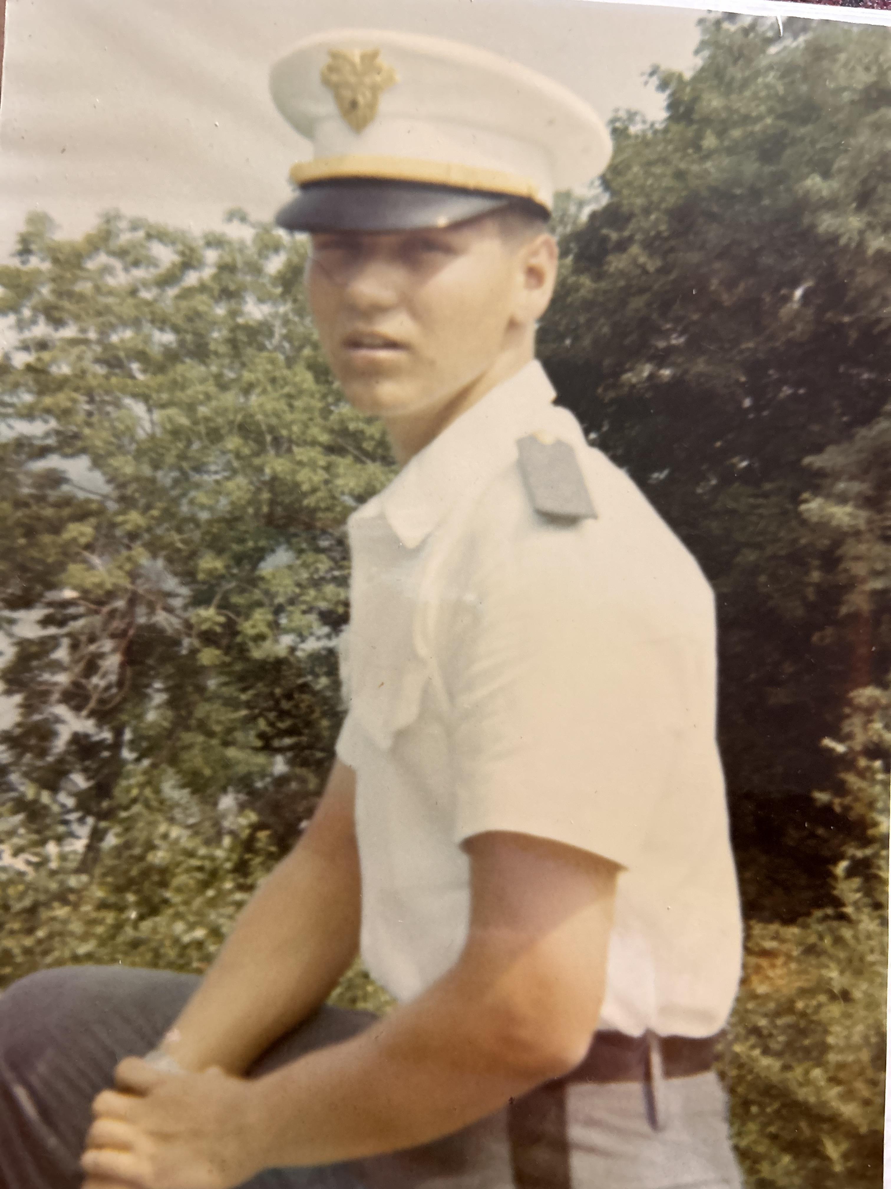 A young soldier sits confidently in uniform, surrounded by greenery, during training.