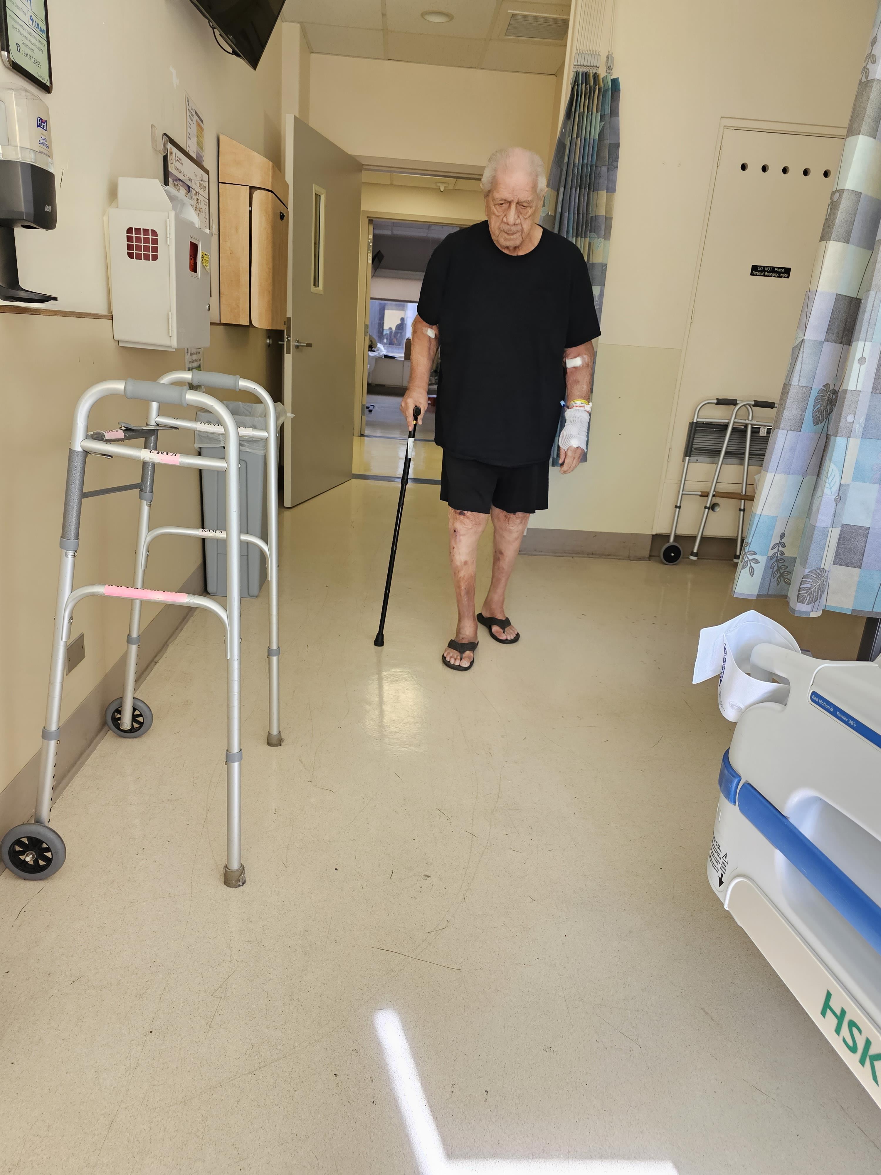 An elderly man uses a cane while walking through a hospital corridor in minimal clothing.