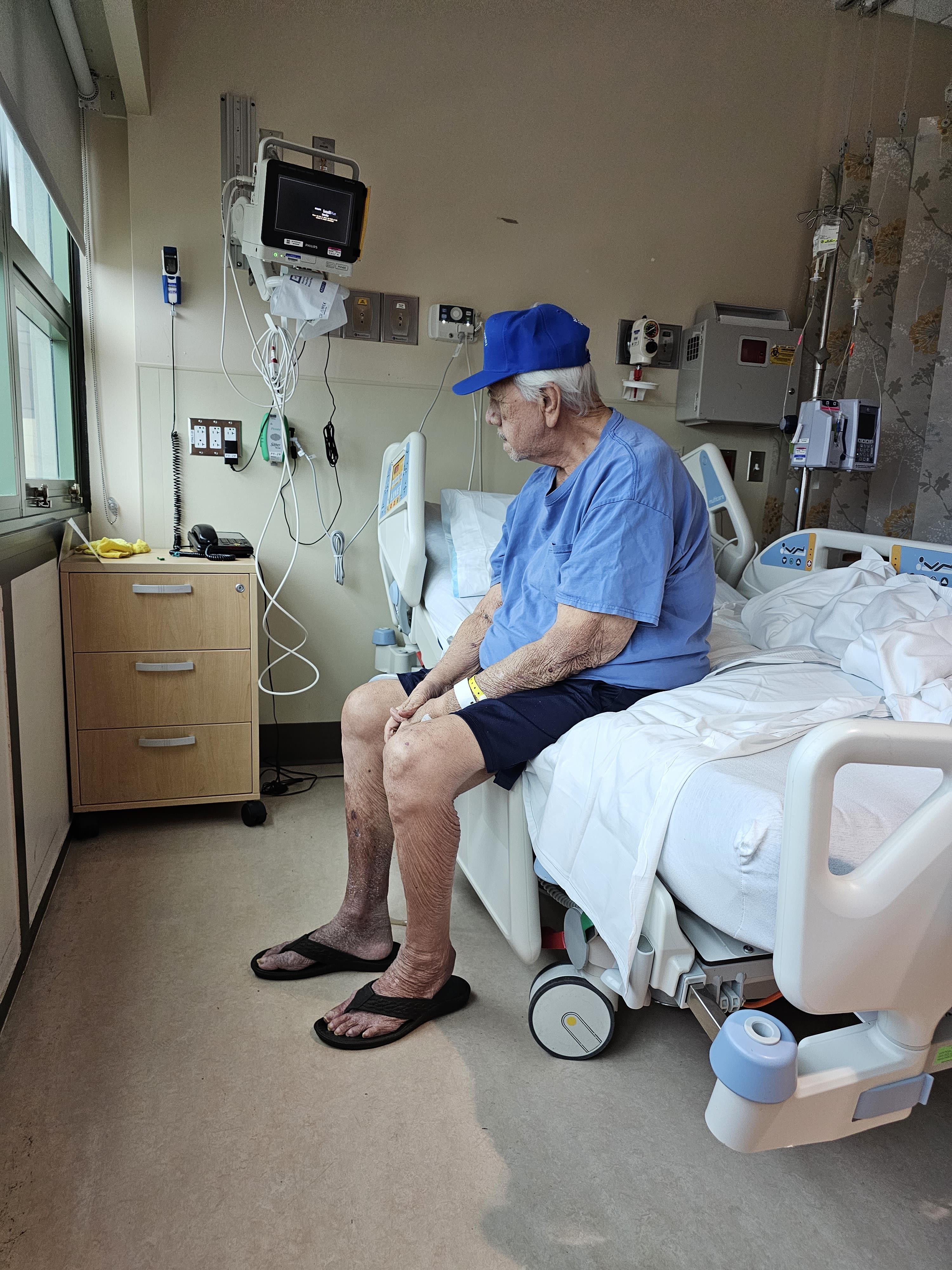 An elderly man in a hospital room sits on the edge of a bed, gazing out the window.