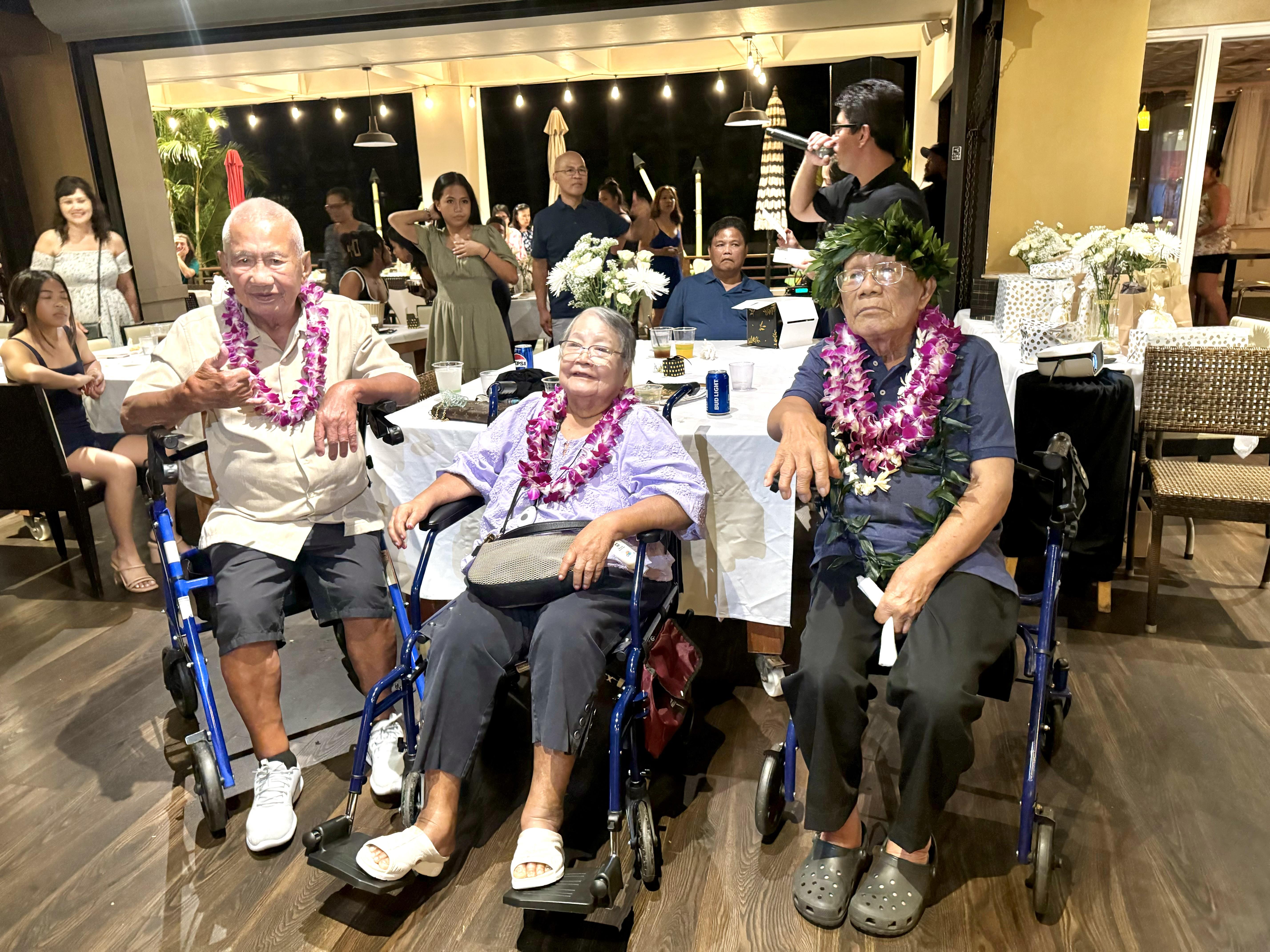 Three elderly individuals smile and interact during a lively celebration surrounded by others.